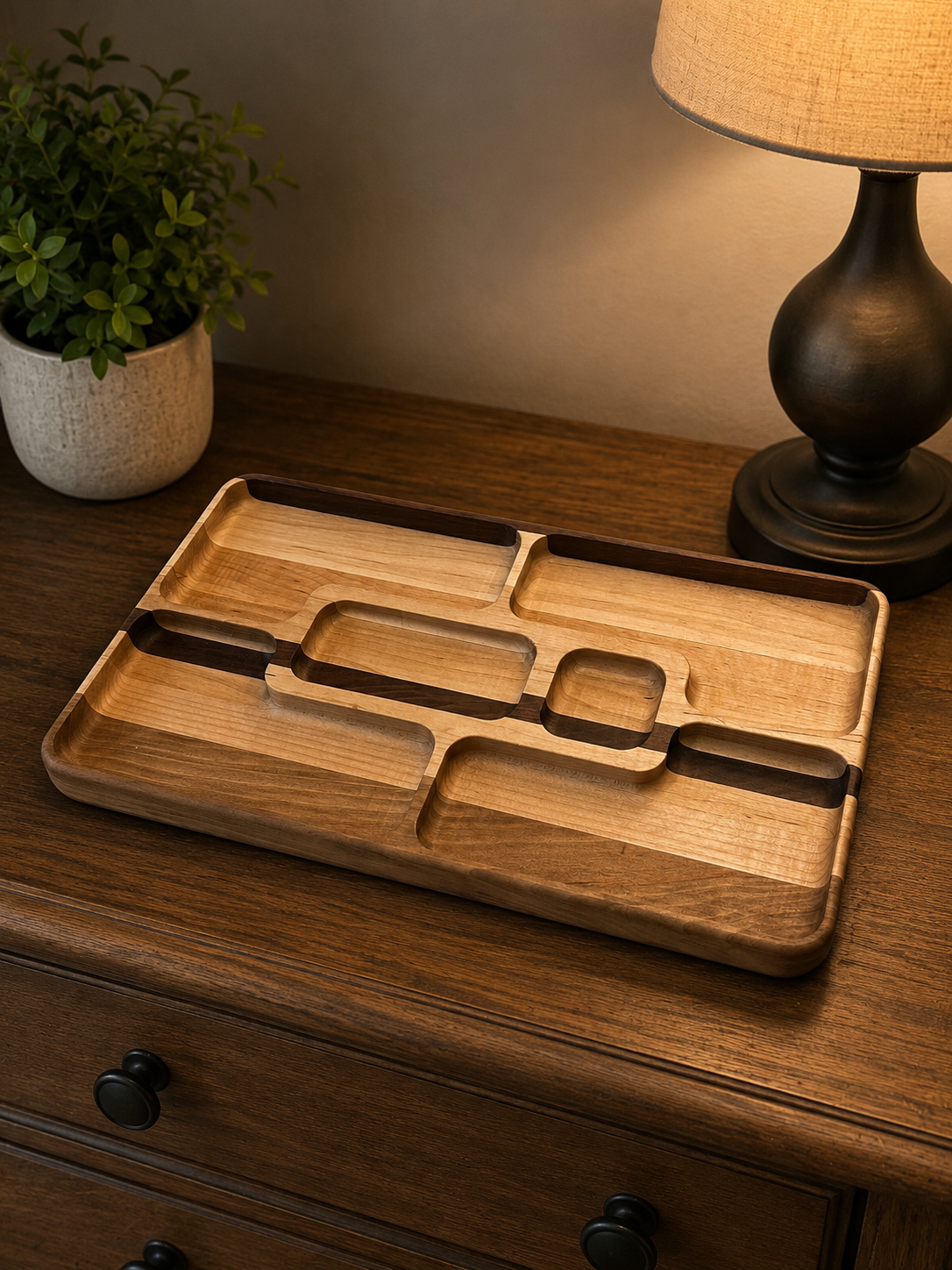 Wooden compartmentalized tray on a dark wooden dresser next to a black table lamp and a potted green plant.