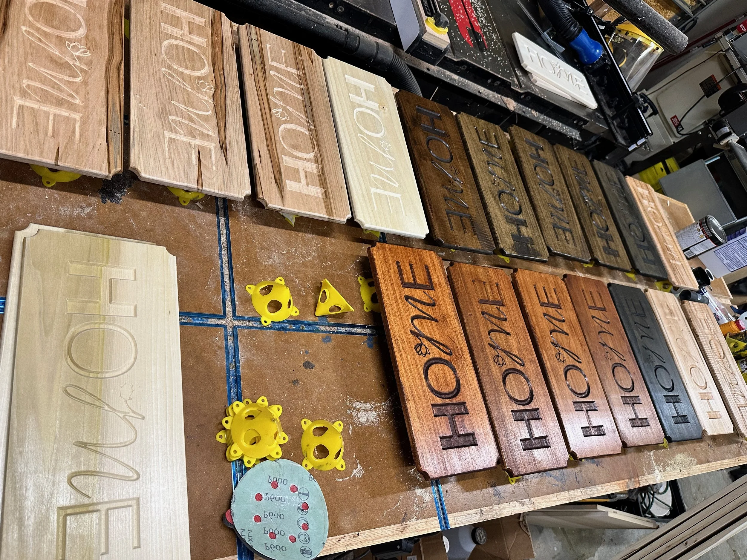 Wooden signs with carved and painted words like 'HOME', 'HAPPY', and 'LOVE' on a workbench in a woodworking shop.