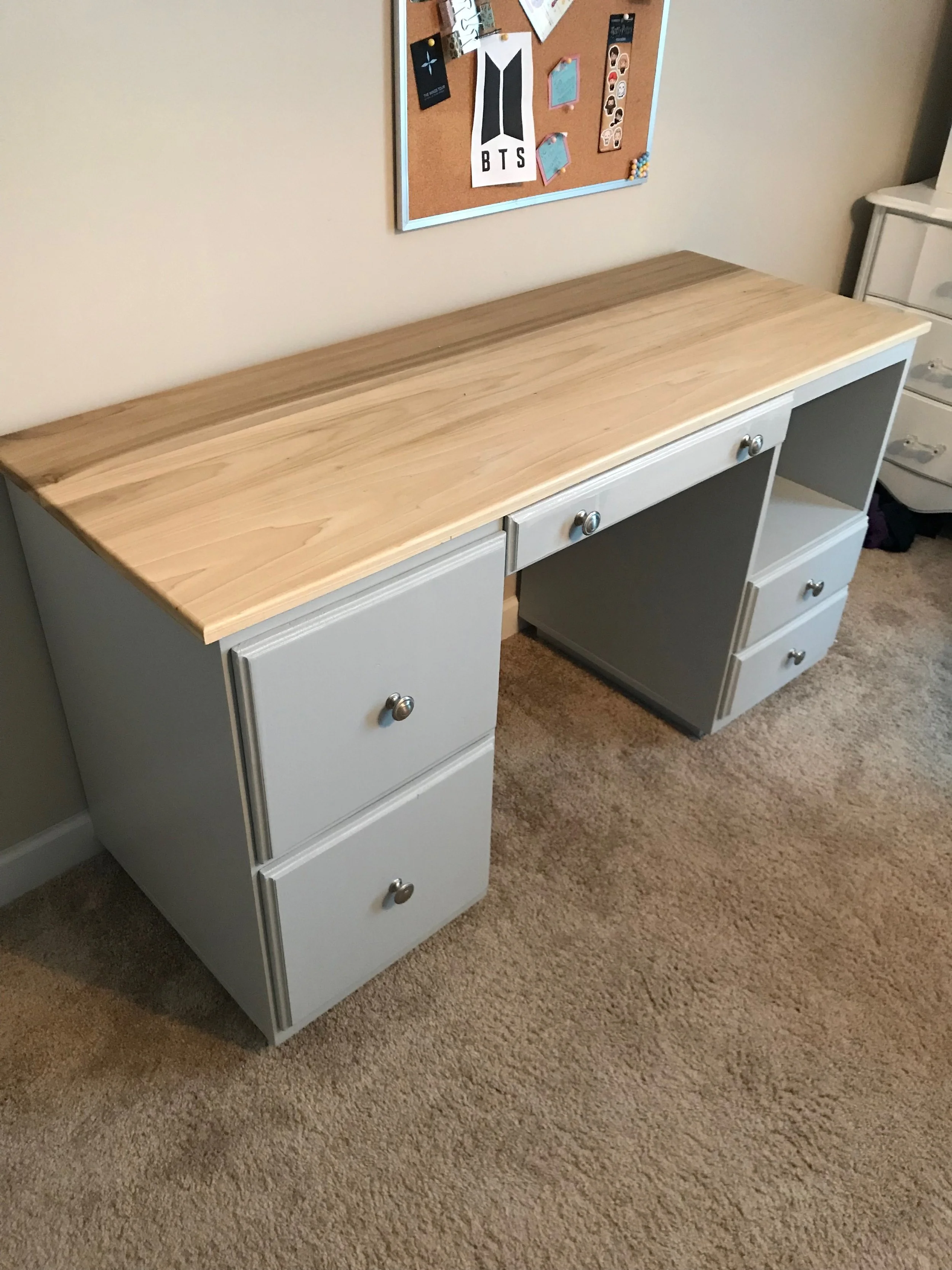 A piece of furniture with a light wood top and gray drawers and doors, situated on a beige carpeted floor, with a bulletin board on the wall above it.