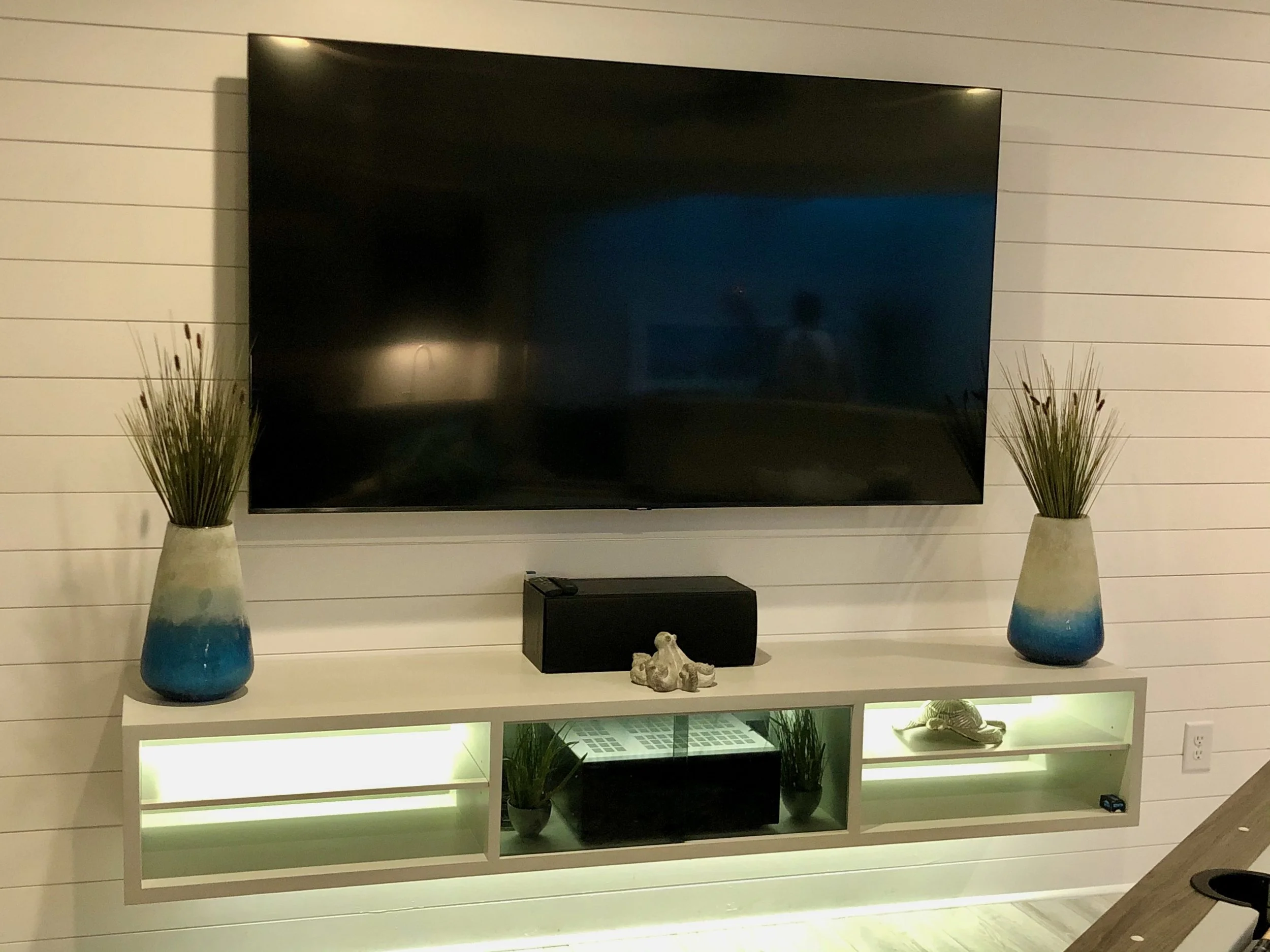 Living room entertainment area with a large flat-screen TV mounted on a white paneled wall. On either side of the TV, there are tall vases with decorative grass. Below the TV, a white shelf holds various decorative items, including a sculpture, small planter, and black electronic devices. The room is well-lit with a modern and minimalist decor style.