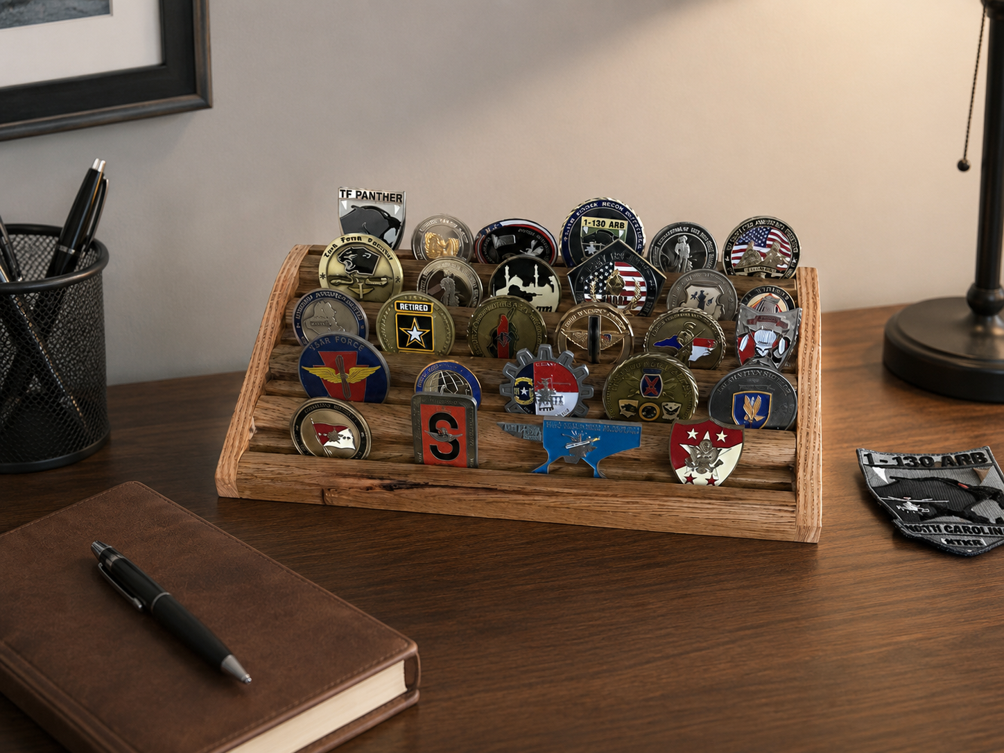 A wooden tray with military patches and medals on a desk, with a black pen, a brown notebook, a black pen holder, and a desk lamp nearby.