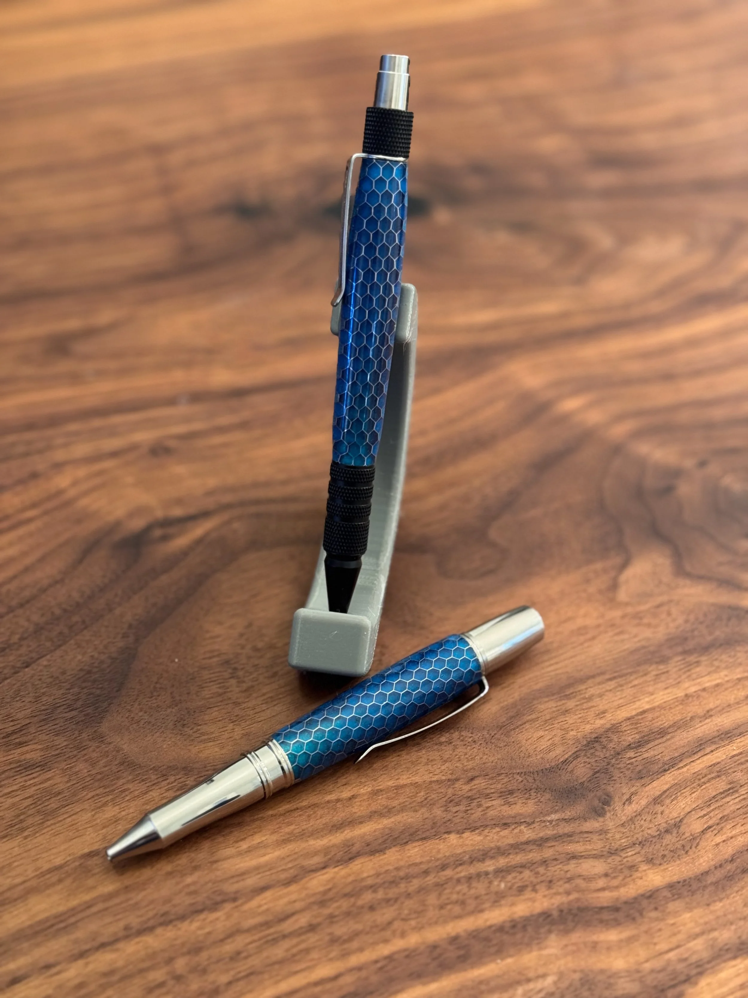 A blue and silver pen with a honeycomb pattern on the barrel rests on a wooden surface. The pen has a silver clip and tip, and a matching blue honeycomb-patterned pen cap lying nearby.