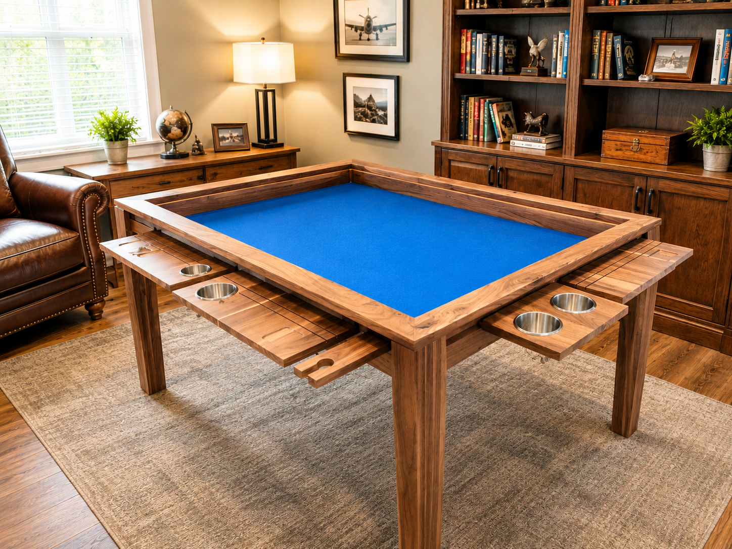 A room with a wooden shuffleboard table with a blue surface, cup holders on the sides, and a wooden extension. There's a leather armchair, a window with blinds, a wooden cabinet with a lamp, picture frames, and books. The room has beige walls, a rug, and decorative items including plants and pictures.