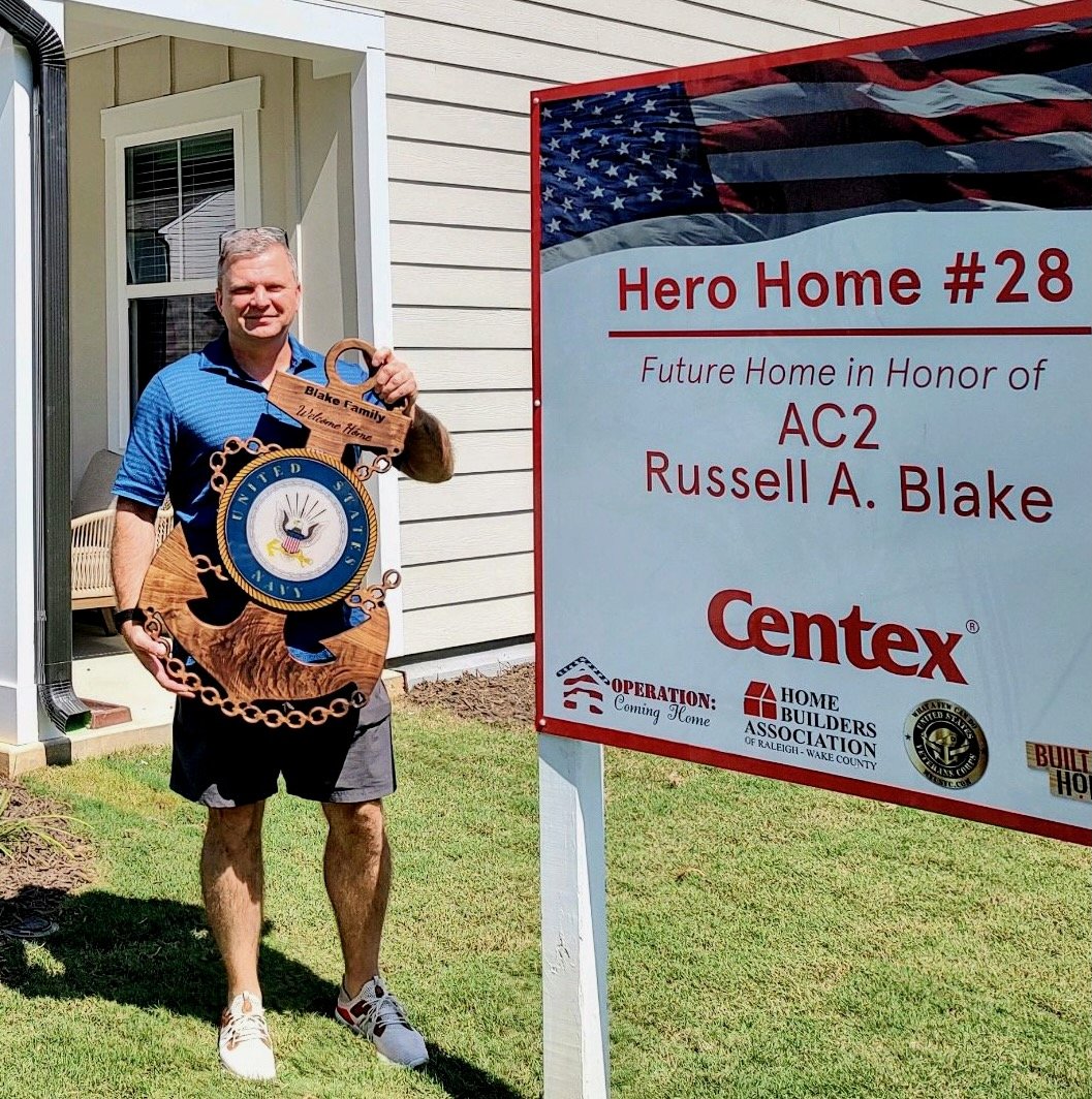 A man standing outside a house holding a large wooden and metal star-shaped sign with the United States emblem, near a sign indicating the future home of Russell A. Blake, part of a community project honoring heroes.