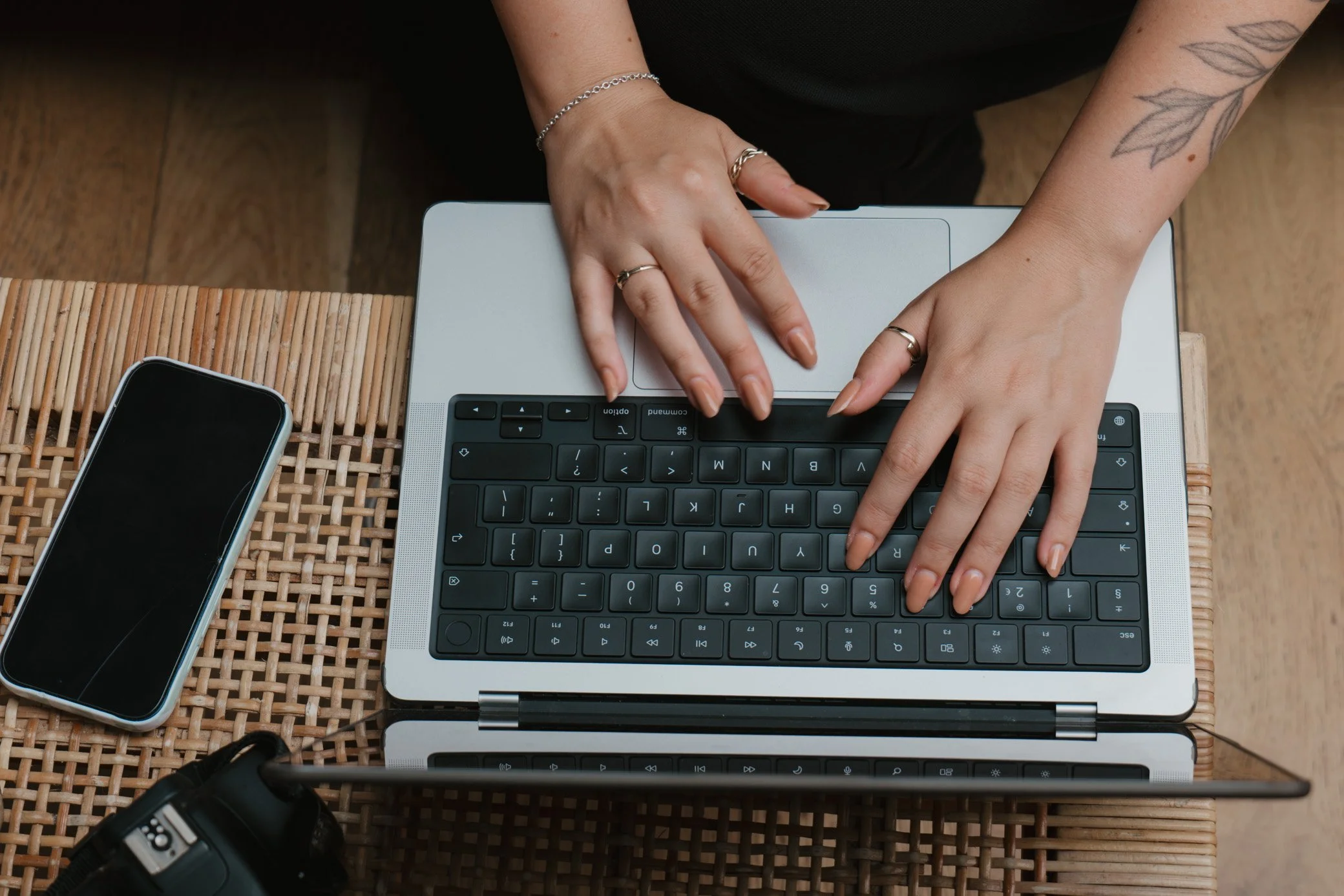 Hands typing on a laptop keyboard with a smartphone and camera on a woven table.