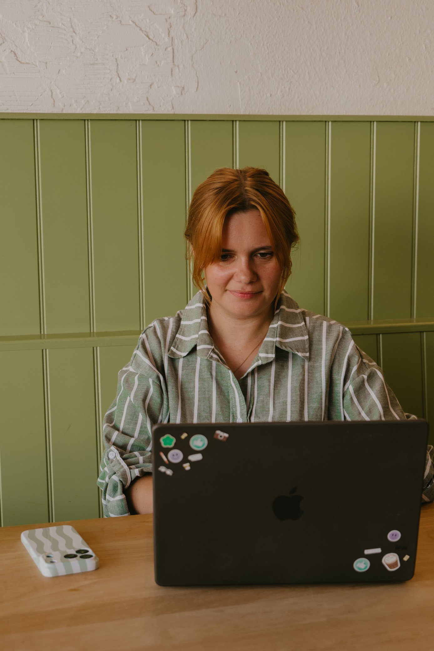 A woman with reddish hair in a striped shirt sitting at a wooden table using a black laptop decorated with stickers, with a green-paneled wall behind her. Leah Croft, social media manager based in Truro, Cornwall