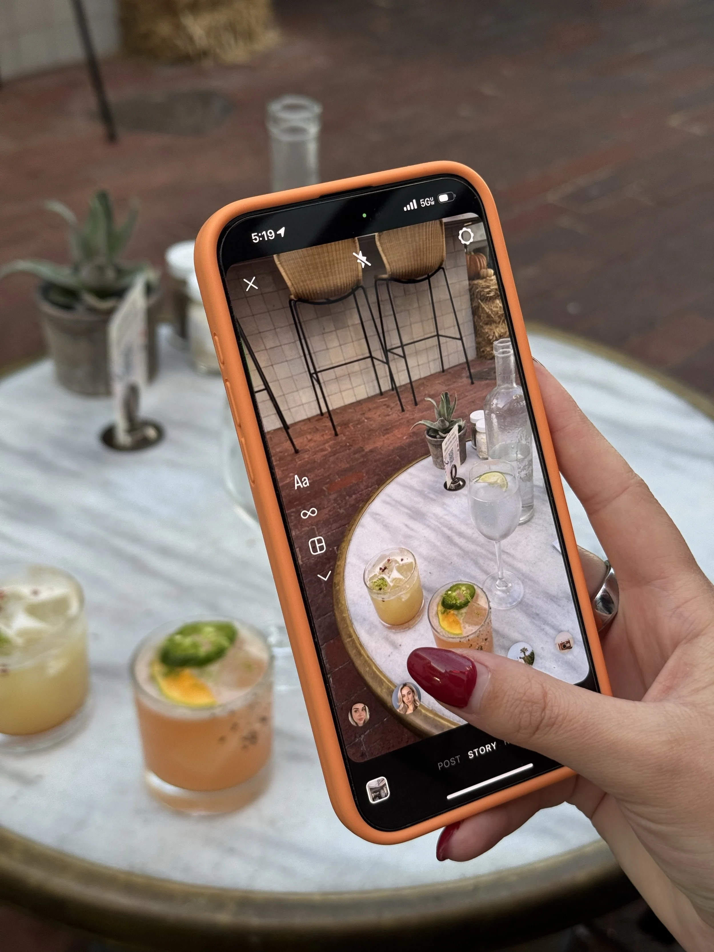 A hand with red nail polish holding an orange phone, taking a photo of a table with several drinks. The drinks are in glasses with slice fruit and mint leaves, set on a white tablecloth with a gold rim. The background shows a table with white tile wall and chairs with wicker seats.