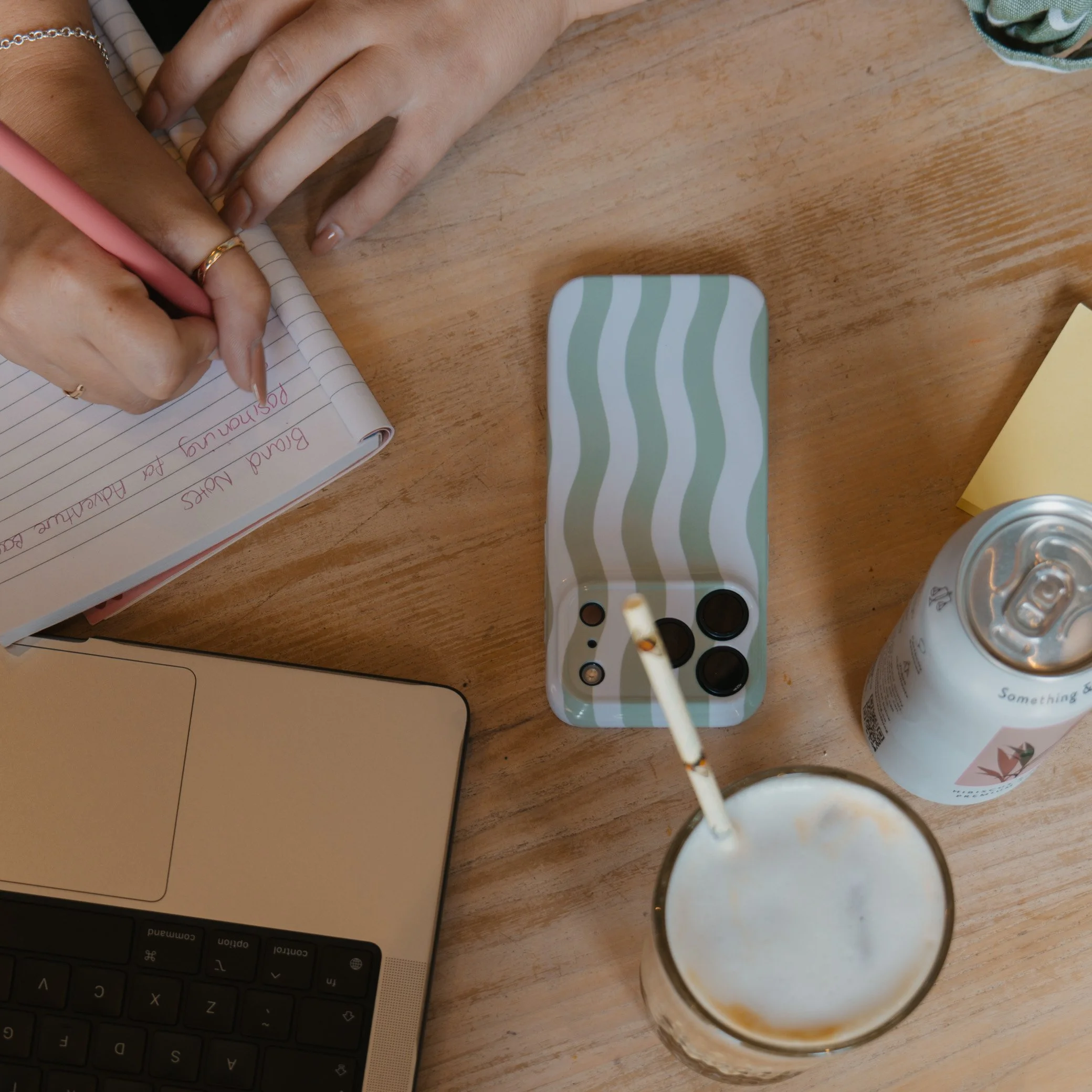 Monthly social media management for Cornwall small businesses. Leah writing in a notebook with a pink pen, on a wooden table alongside a closed laptop, a striped rectangular container, a computer keyboard, a drink in a glass with a straw, and a can.