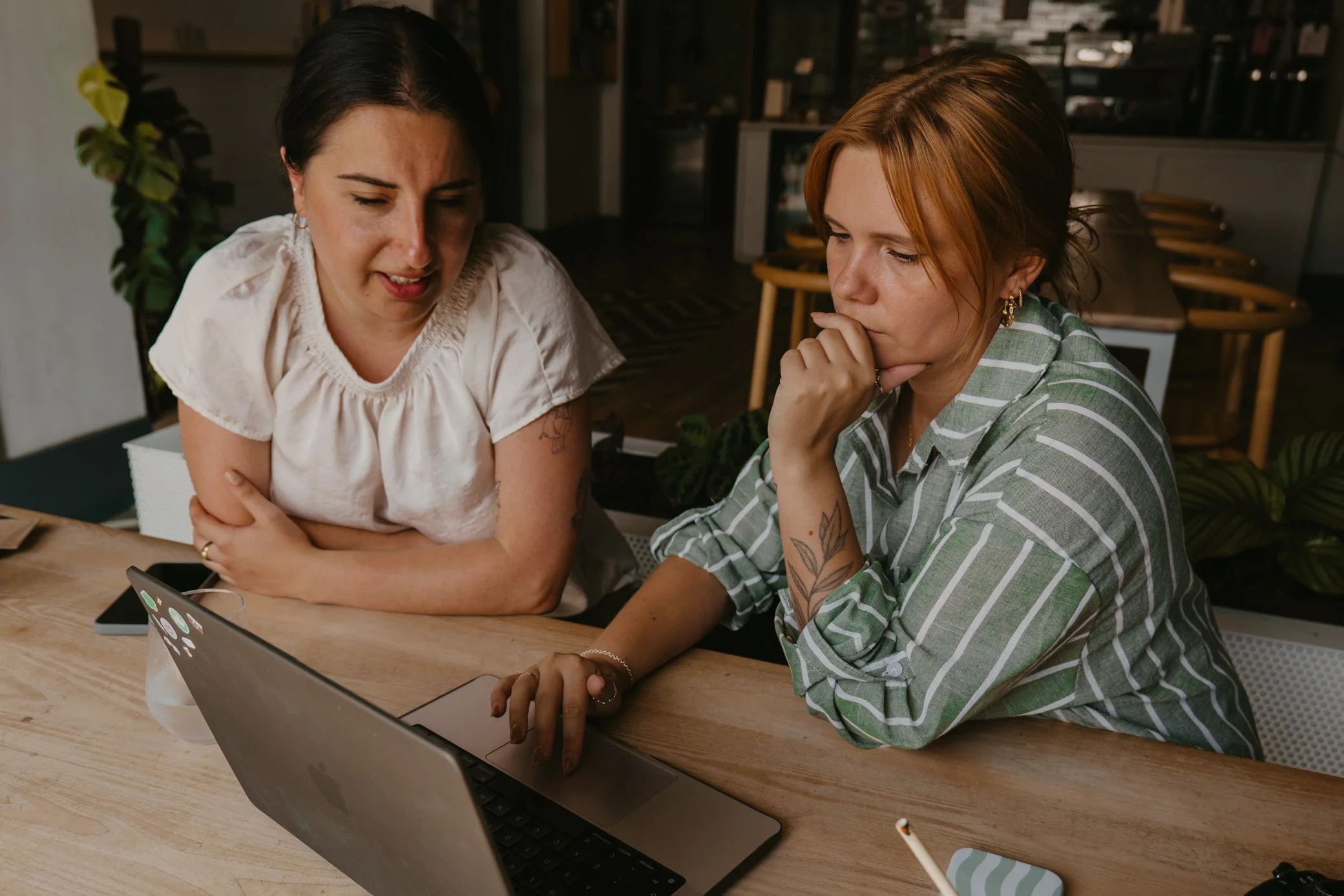 Two women seated at a wooden table working on a laptop in an indoor setting. One woman with dark hair and a white blouse, the other with auburn hair, striped shirt, and tattoos on her arm, intently looking at the laptop screen.
