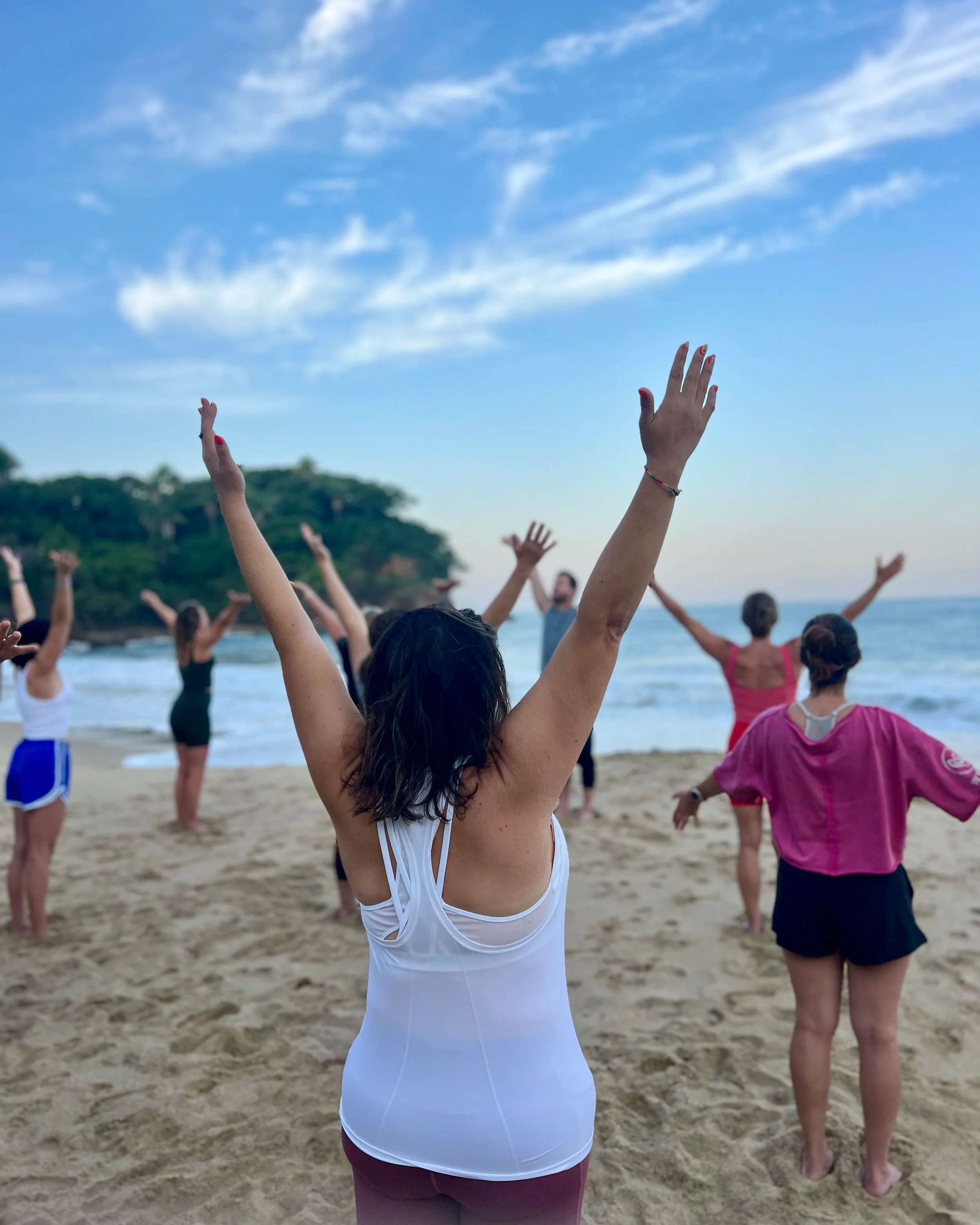 A group of people practicing yoga on the beach during the daytime, with arms raised towards the sky and a blue sky with clouds in the background.