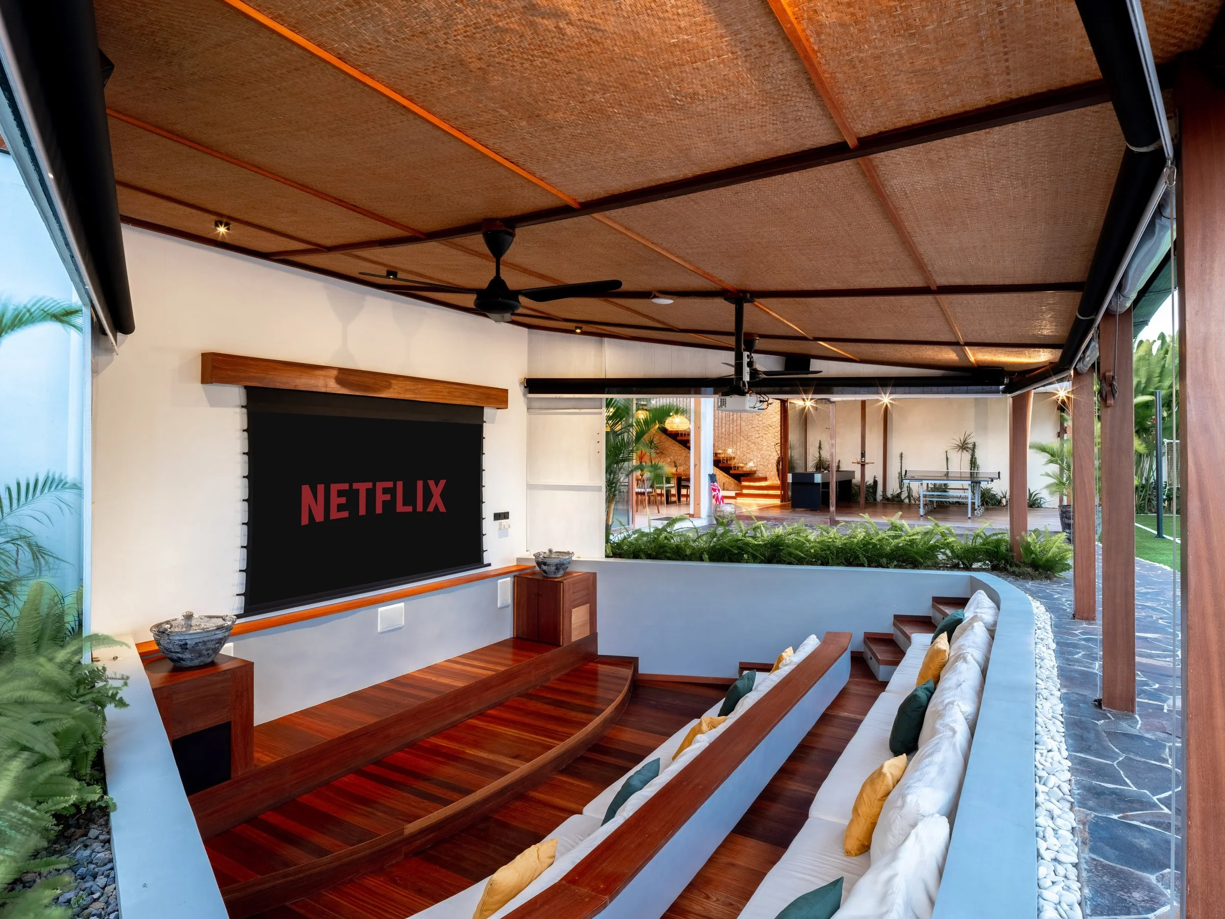 Indoor lounge area with a wooden ceiling, black ceiling fan, white walls, a large NETFLIX screen, and wooden furniture with cushions.