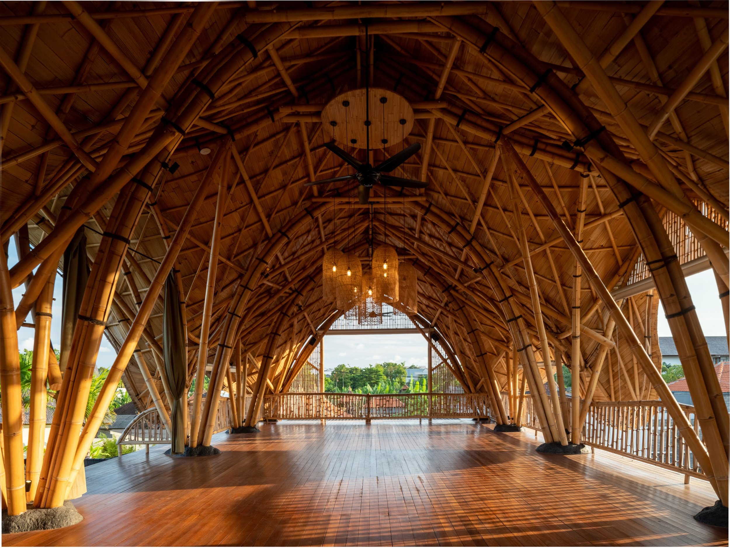 View of an elegant indoor space with bamboo architecture, high vaulted bamboo ceiling, large hanging light fixtures, wooden flooring, and large windows showing lush green landscape outside.