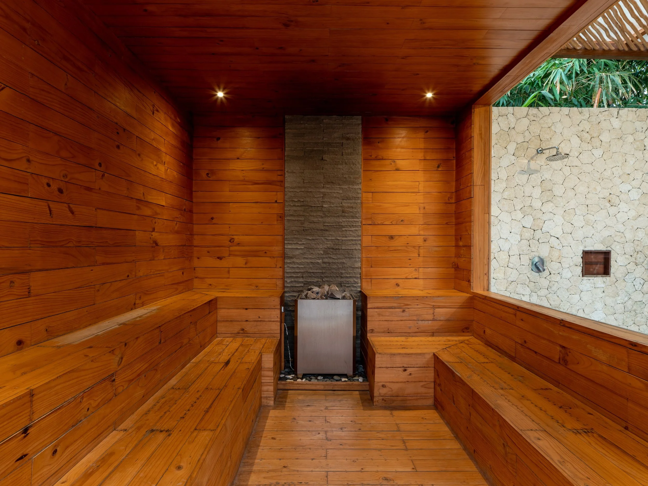 Outdoor shower area with wooden walls and ceiling, stone wall with showerhead, and greenery visible outside.