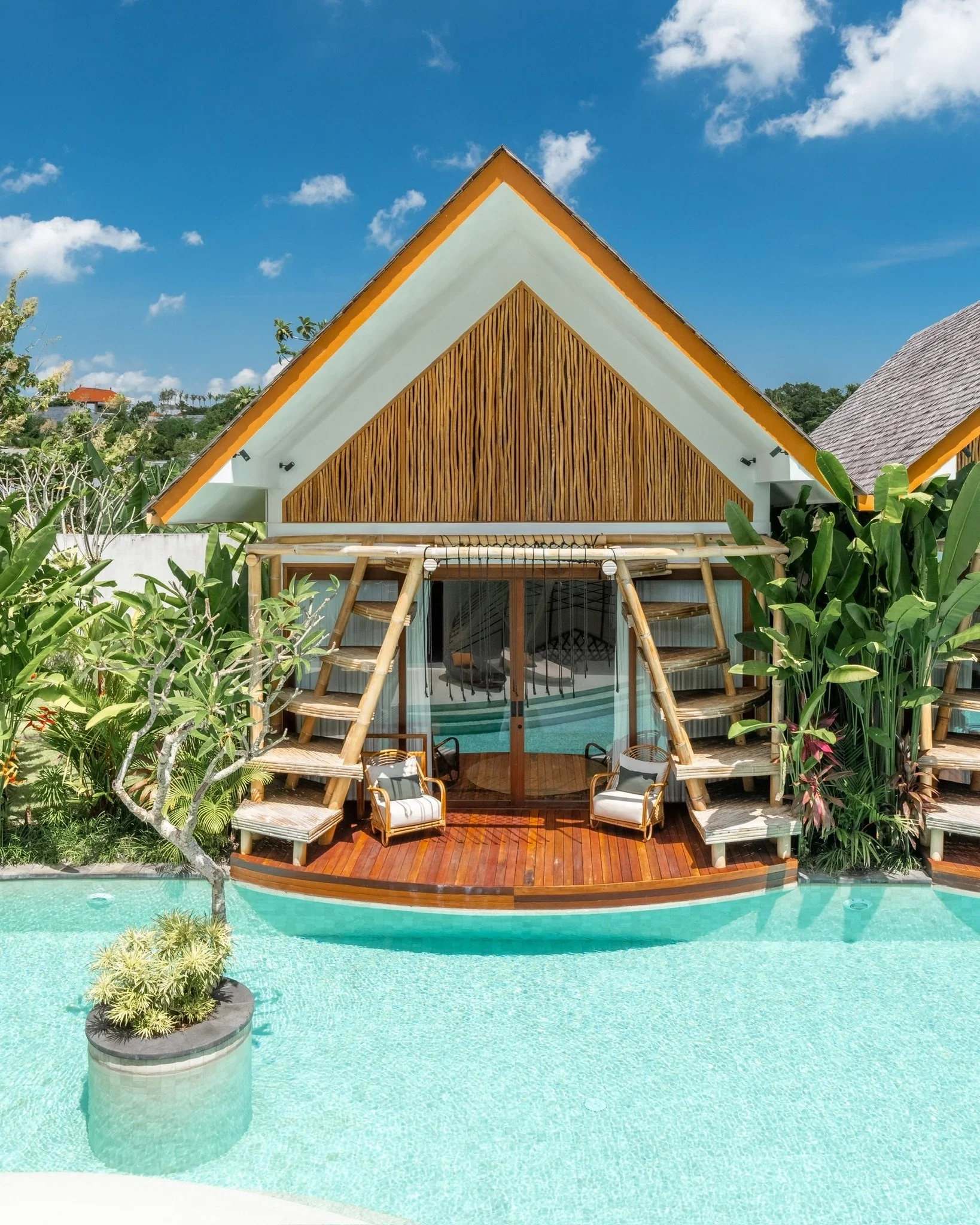 A modern house with a steep roof, wooden paneling, and a glass door, surrounded by lush greenery and a swimming pool with clear water.
