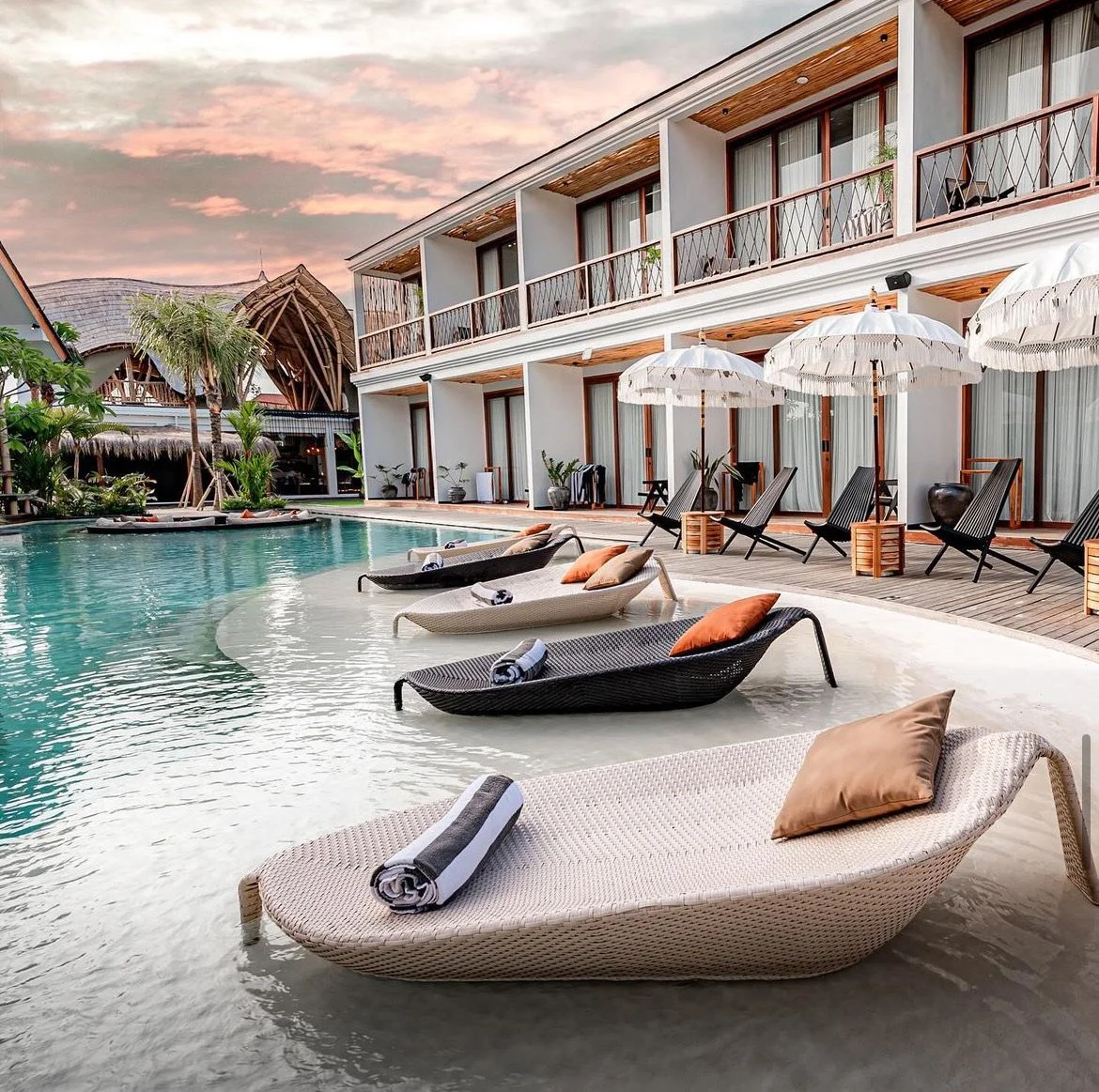 Luxury hotel pool area with lounge chairs, umbrellas, and a modern building with balconies, tropical plants, and a sunset sky.