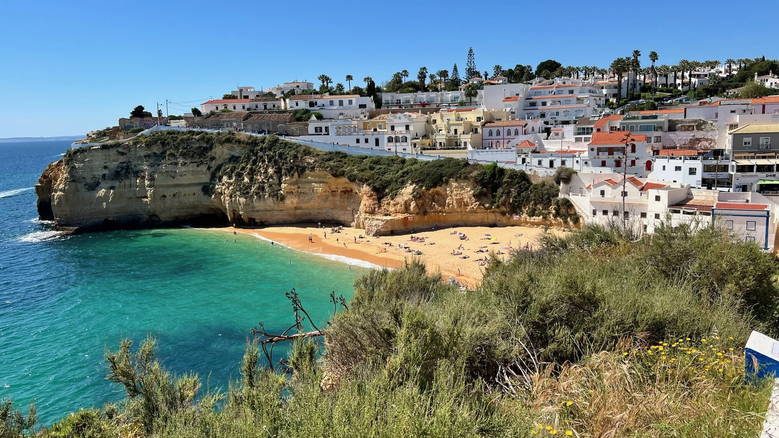 Scenic view of a coastal town with white buildings with red-tiled roofs perched on cliffs. A small sandy beach with people and umbrellas is visible below, with turquoise ocean water lapping at the shore. Green shrubs and grasses frame the foreground.