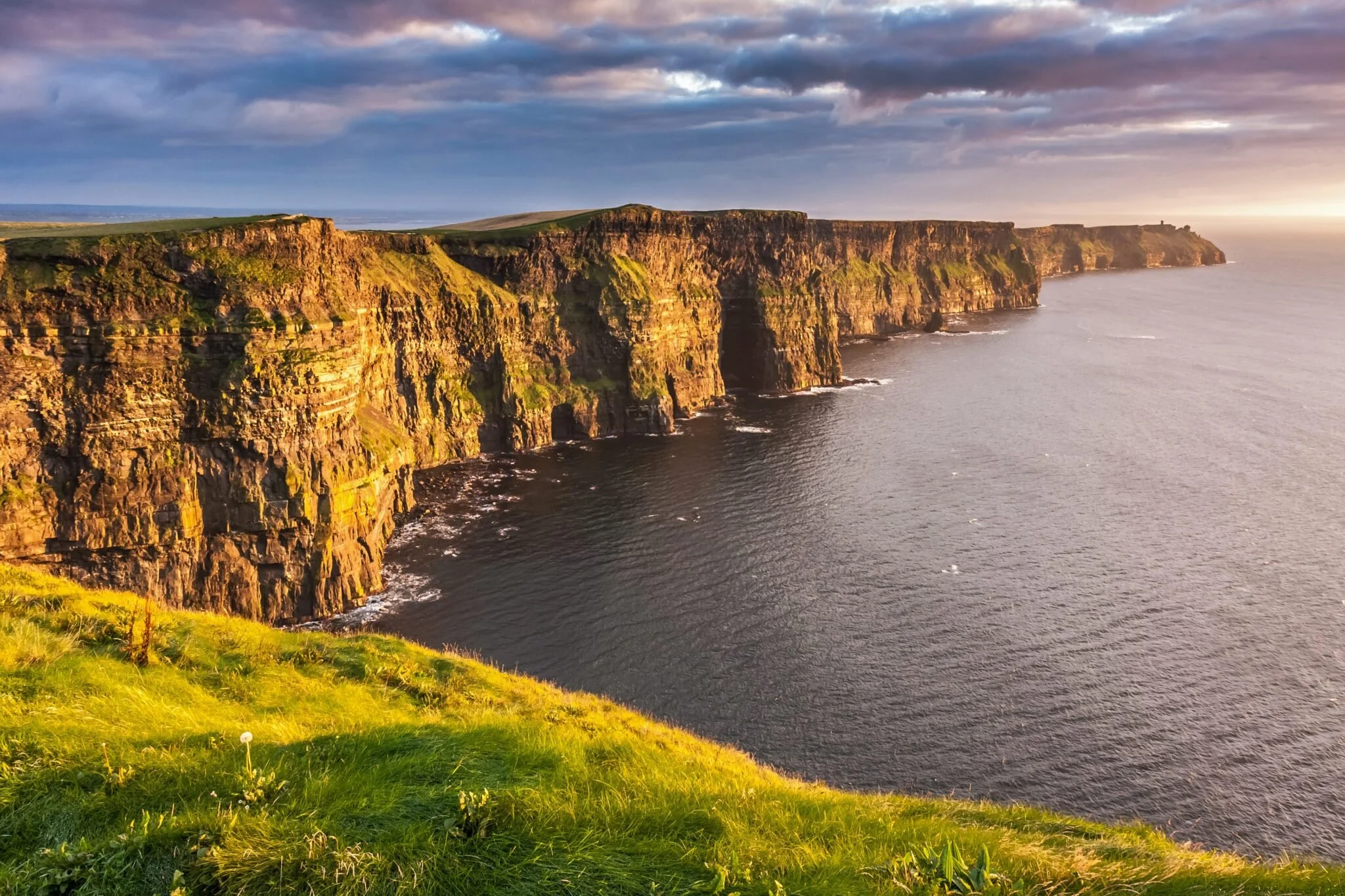 Cliffs along the ocean during sunset with cloudy sky and green grass in the foreground.