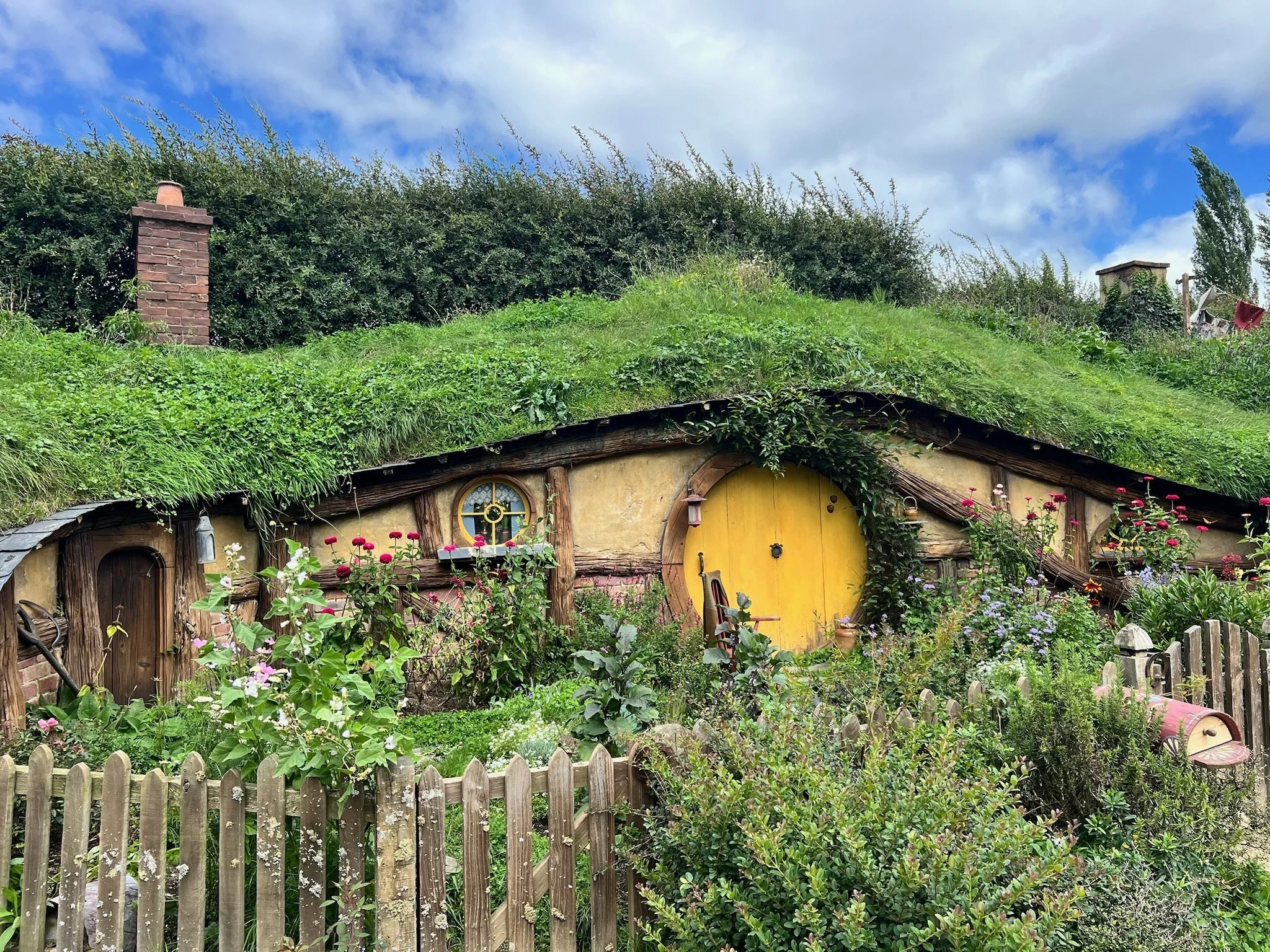 A charming cottage with a yellow door, surrounded by colorful flowers and garden, with a green, grassy roof and brick chimneys under a partly cloudy sky.