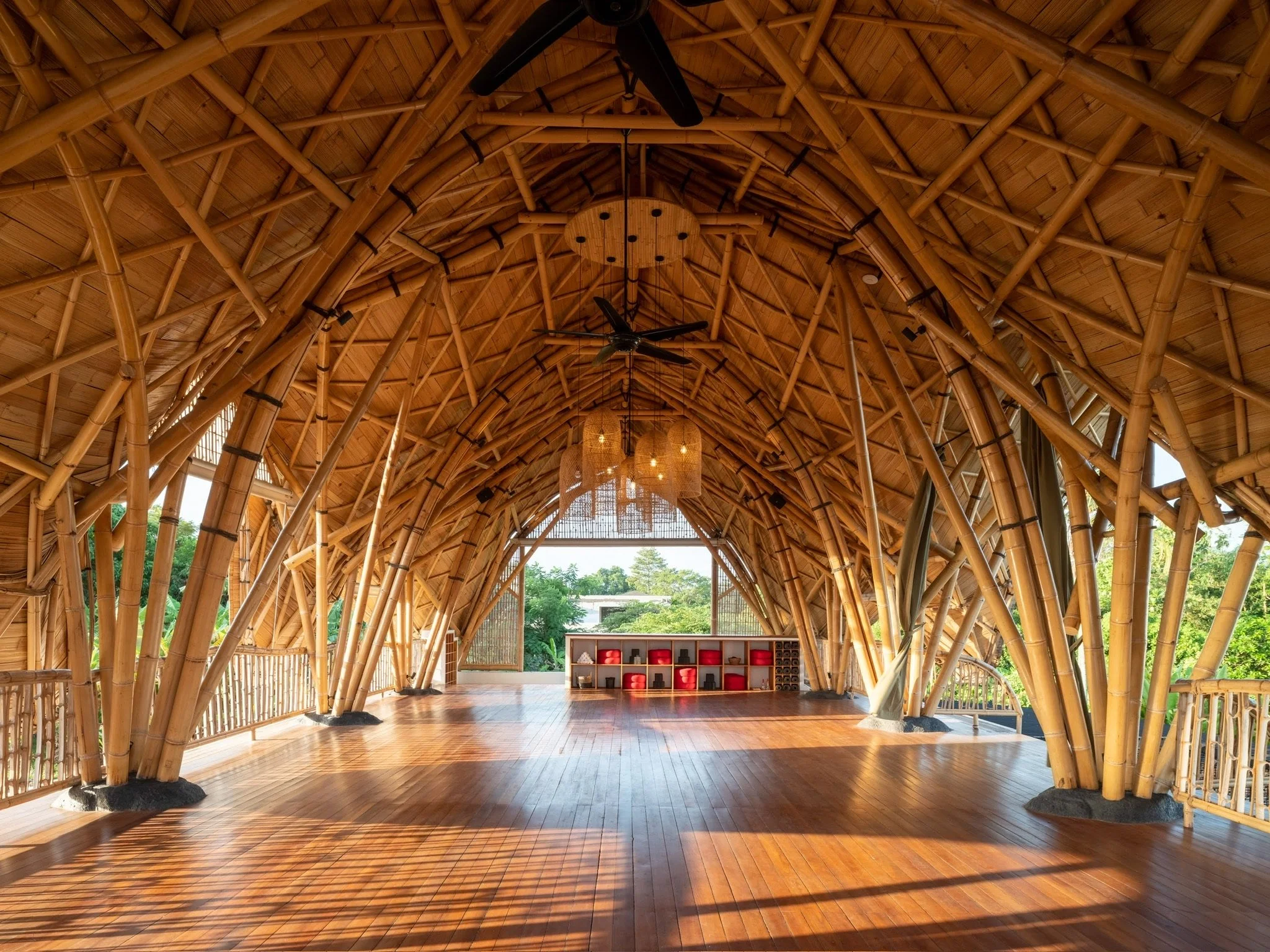 Interior of a building with a bamboo frame ceiling, wooden floor, ceiling fans, hanging lights, racks, and natural light coming through windows.