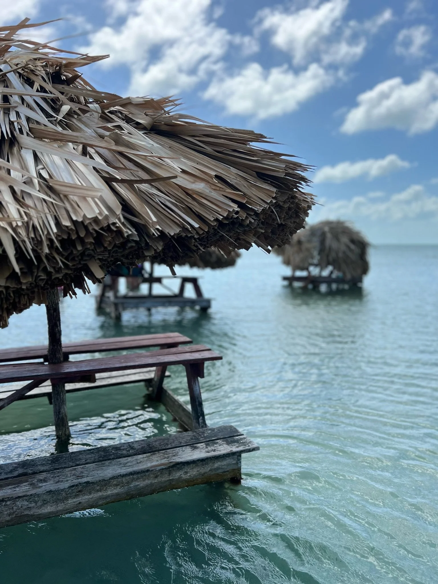 A thatched-roof hut on stilts over ocean water with a clear blue sky and scattered clouds in the background.