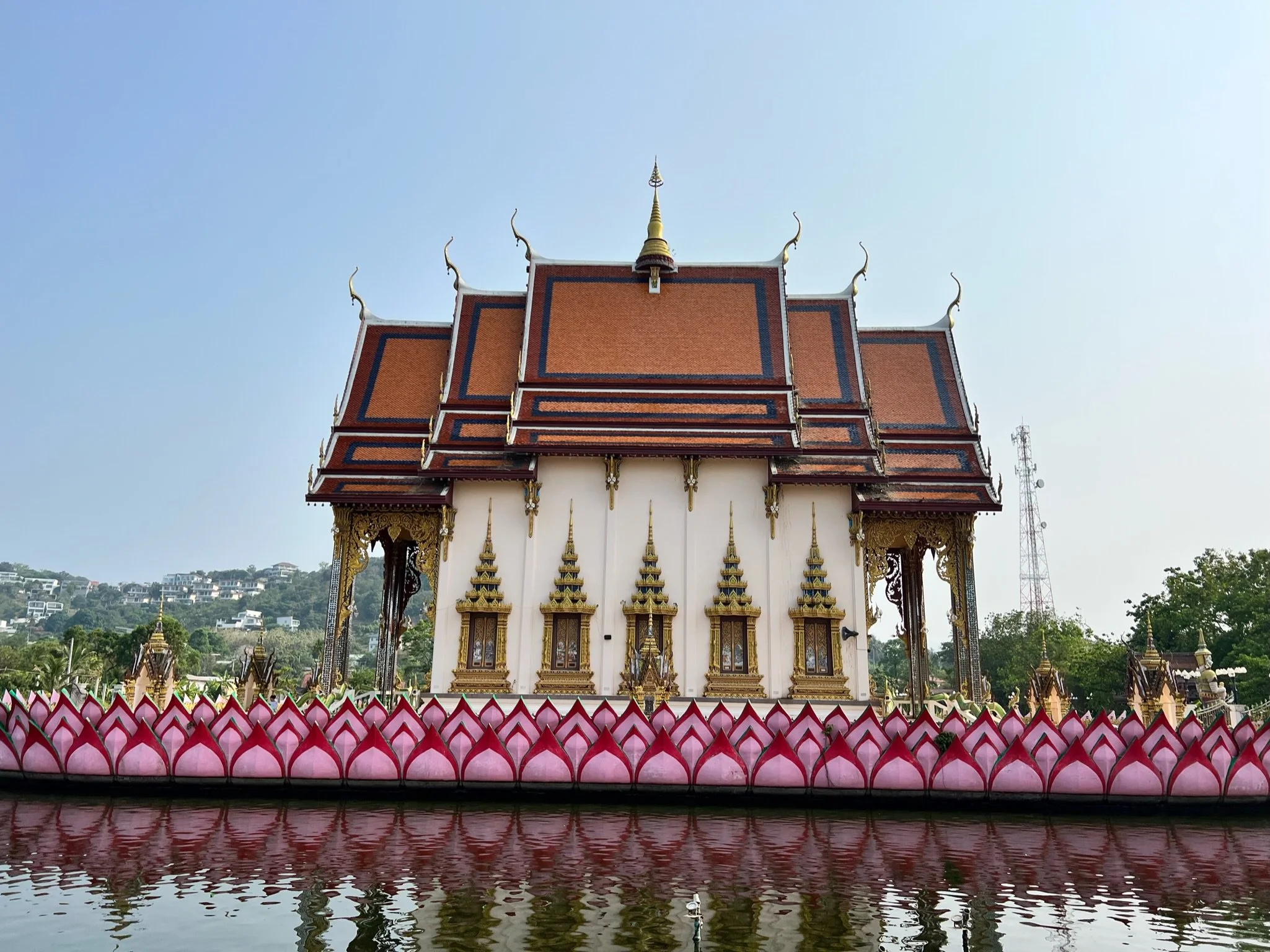 A traditional Thai temple with ornate architectural details, situated near a water body that reflects part of the temple, under a clear blue sky.