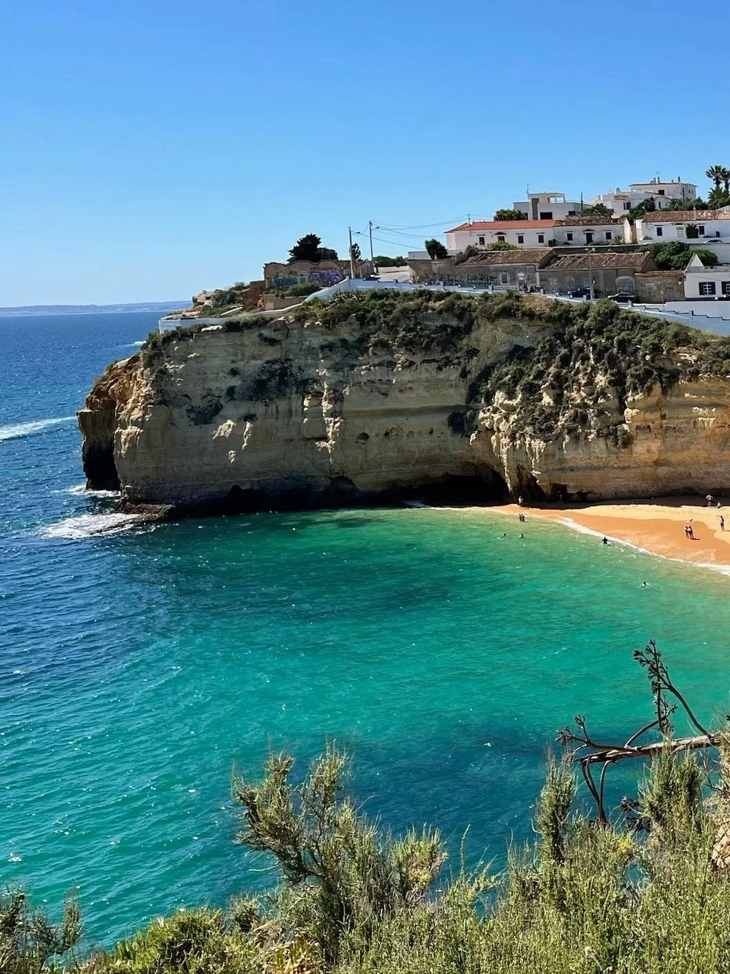 Cliffs overlooking a beach with turquoise water, white houses on the hill, under a clear blue sky.