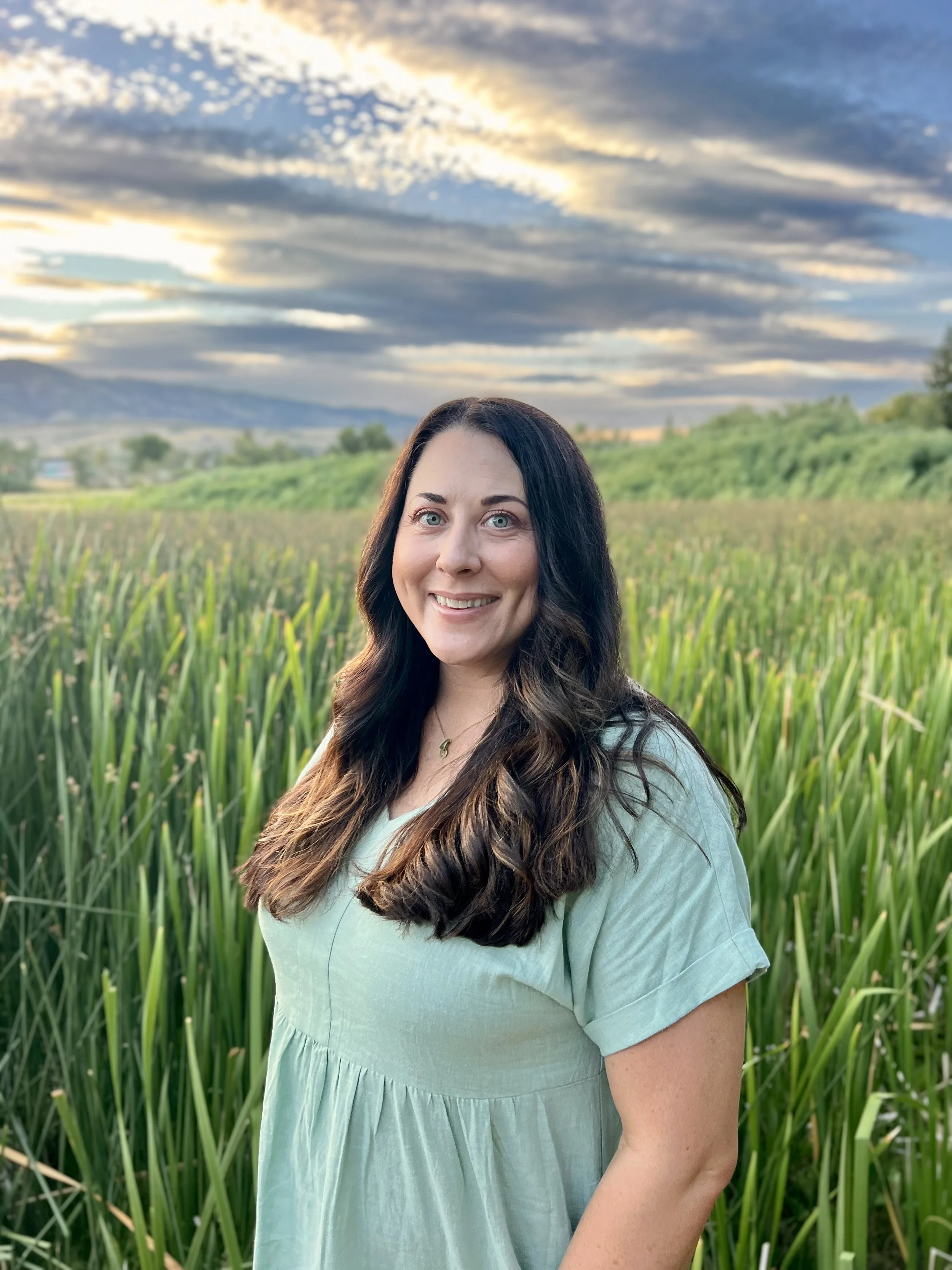A woman with long, wavy brown hair and blue eyes smiling outdoors in a green field with tall grass, with mountains and a partly cloudy sky in the background during sunset.