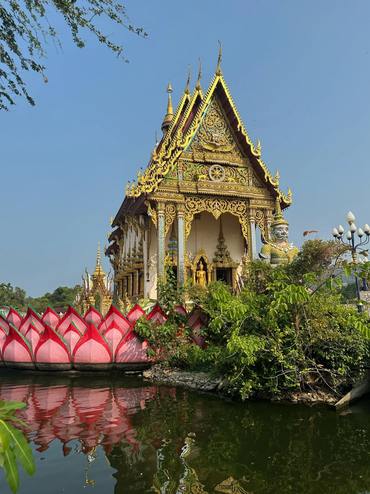 Ornate Thai temple building with decorative golden roof, situated by a body of water with reflections, greenery, and a stone lion statue nearby.