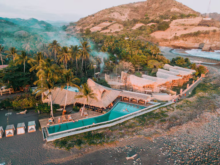 A seaside resort with multiple swimming pools surrounded by palm trees, thatched roofs, and lounge chairs, set against a lush green hillside and ocean.