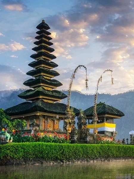 A traditional Balinese temple with multiple tiered pagoda-style roofs, set against a mountainous backdrop with a cloudy sky.