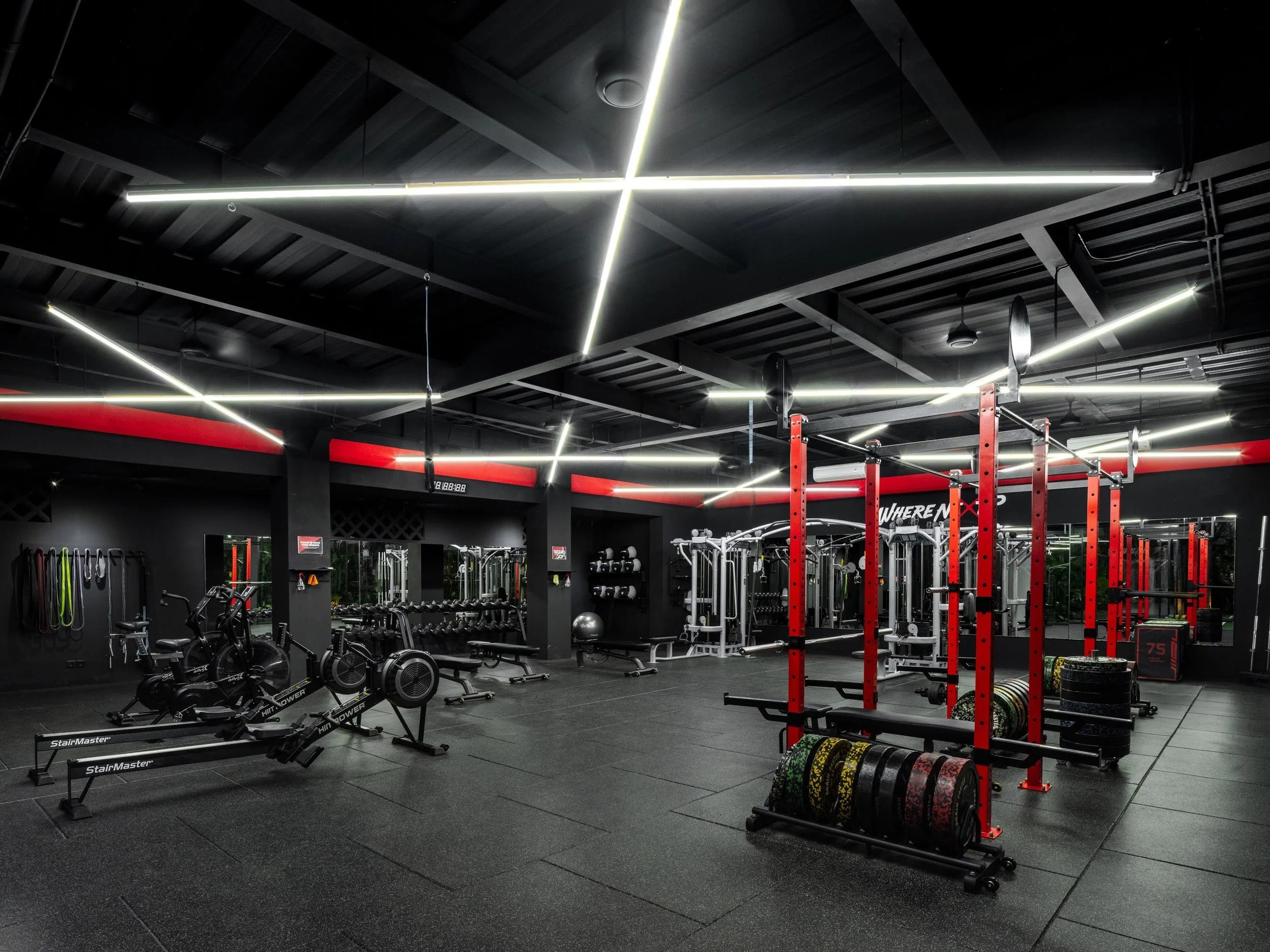 A gym with black walls, ceiling, and floor illuminated by modern LED strip lights. The gym features red squat racks, various weightlifting equipment, rowing machines, and black rubber weights and mats.