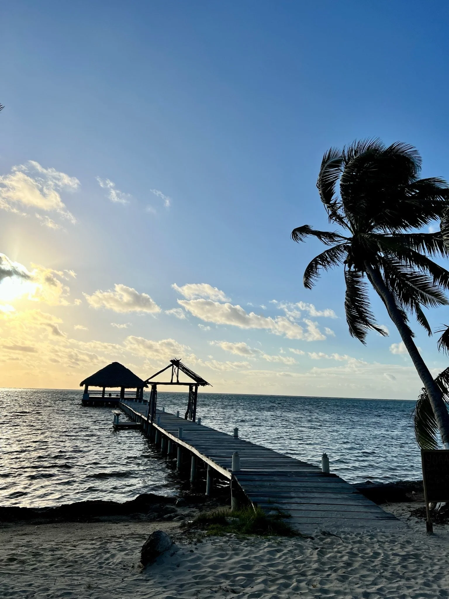 Beach scene at sunset with a wooden pier extending into the ocean, a thatched-roof hut on the pier, a palm tree to the right, and clouds in the sky.