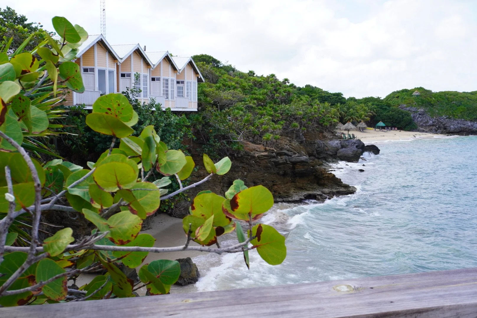 Beachside cottages with green vegetation, rocky shoreline, sandy beach with umbrellas, and ocean waves.
