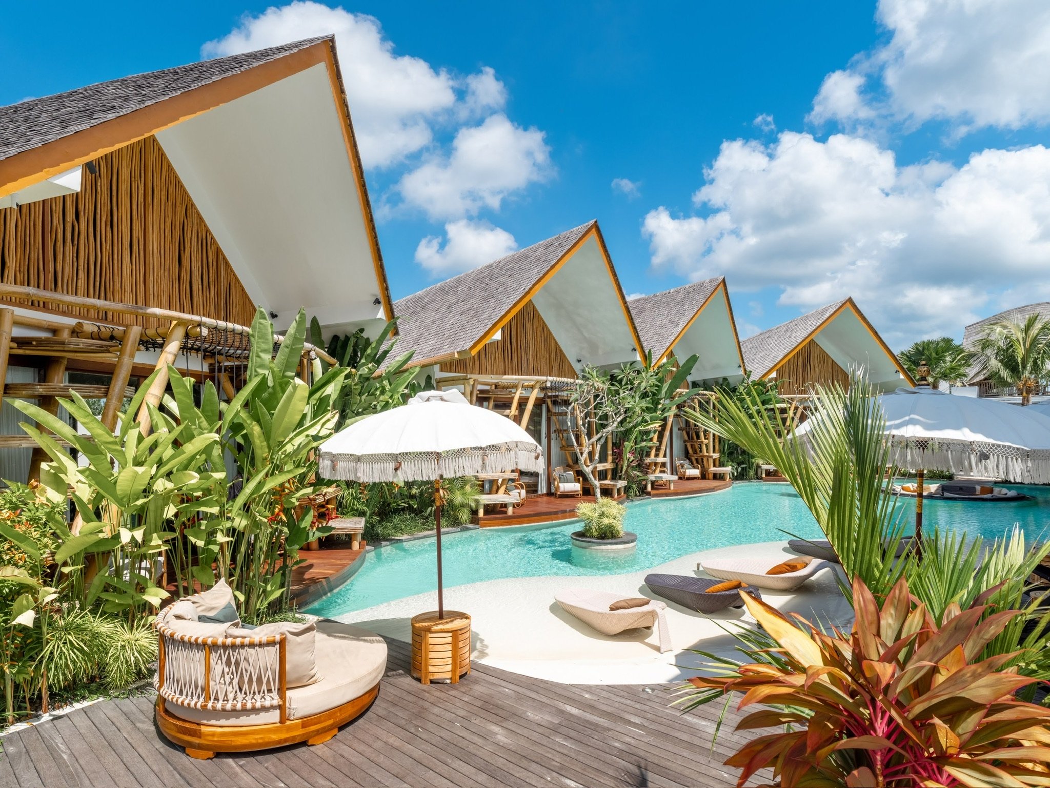 Resort pool area with lounge chairs, umbrellas, lush greenery, and modern cabanas under a blue sky with clouds.