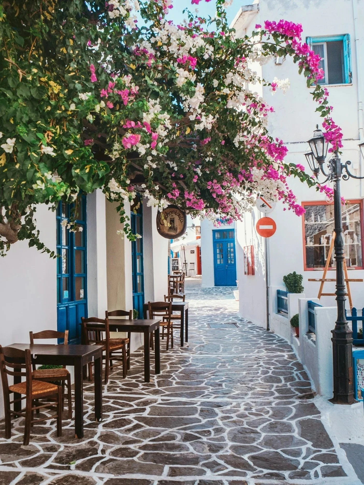 A white-washed sidewalk cafe with wooden tables and chairs, decorated with blue window frames, beneath a blooming bougainvillaea tree on a narrow street in a Mediterranean village.