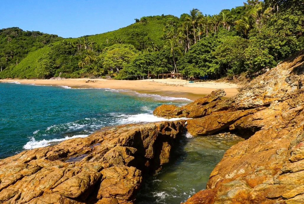A tropical beach with stones in the foreground, clear blue water, a sandy shoreline, and green lush hills with palm trees in the background.