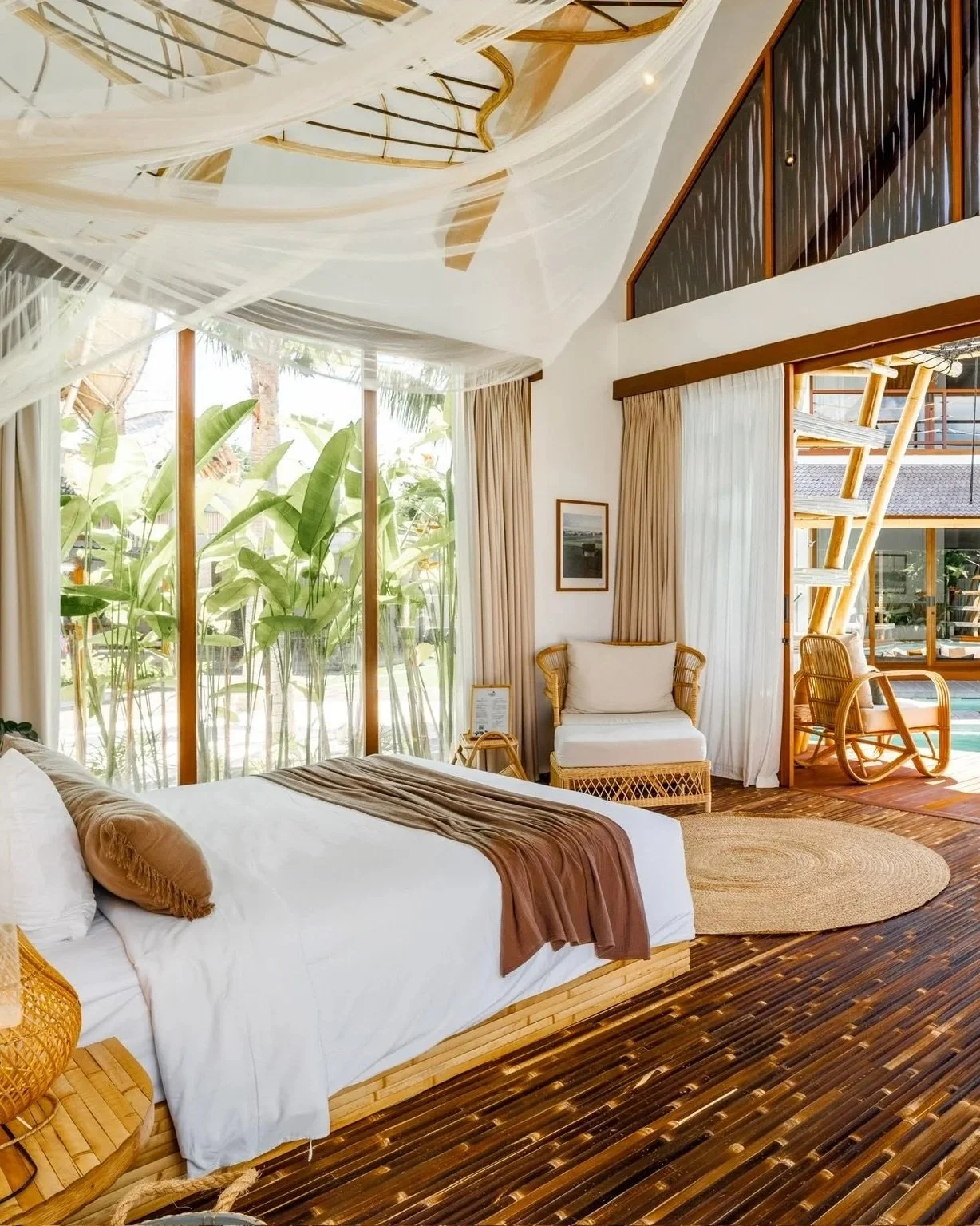 A bright tropical-style hotel room with large glass windows showing lush green plants outside. The room features a white bed with brown accents, rattan furniture, and wooden flooring. There are beige curtains, a framed picture on the wall, and a cozy seating area with a white cushioned chair and bamboo rocking chair on a deck outside.
