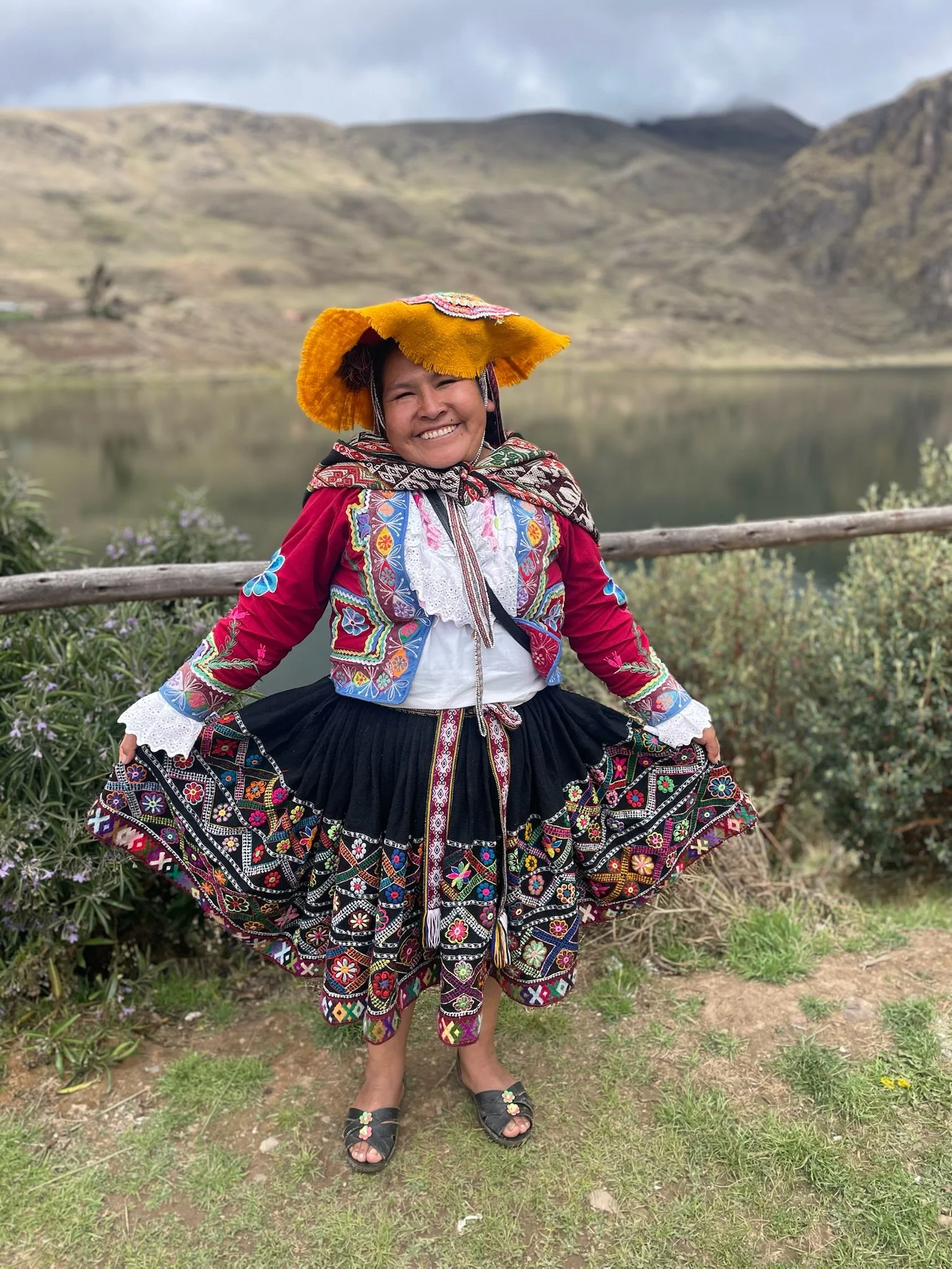 A woman in traditional colorful Peruvian attire, smiling, standing outdoors near a lake with mountainous landscape in background.