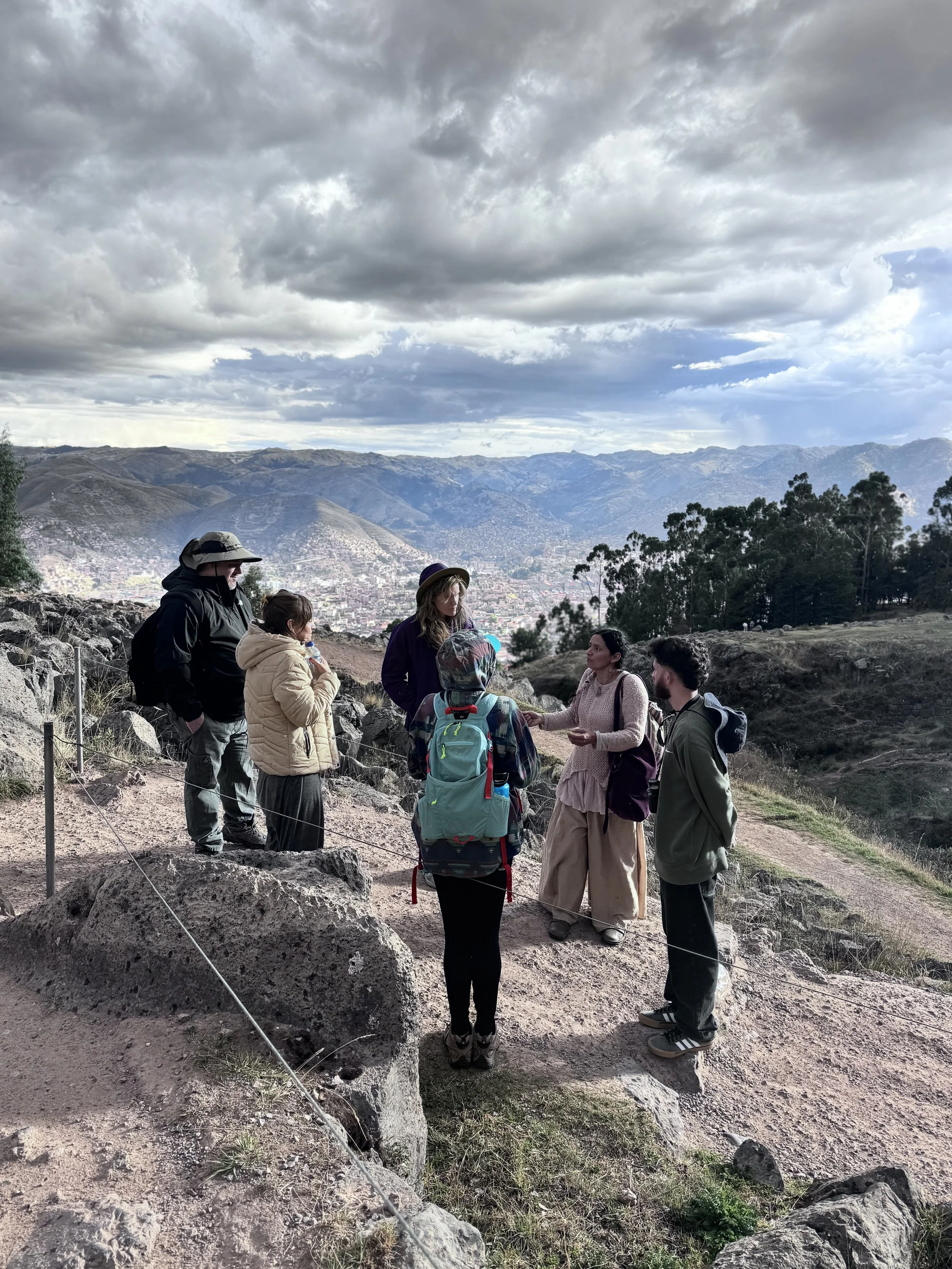 A group of people outdoors on a rocky hillside with a city and mountains in the background, under a cloudy sky.