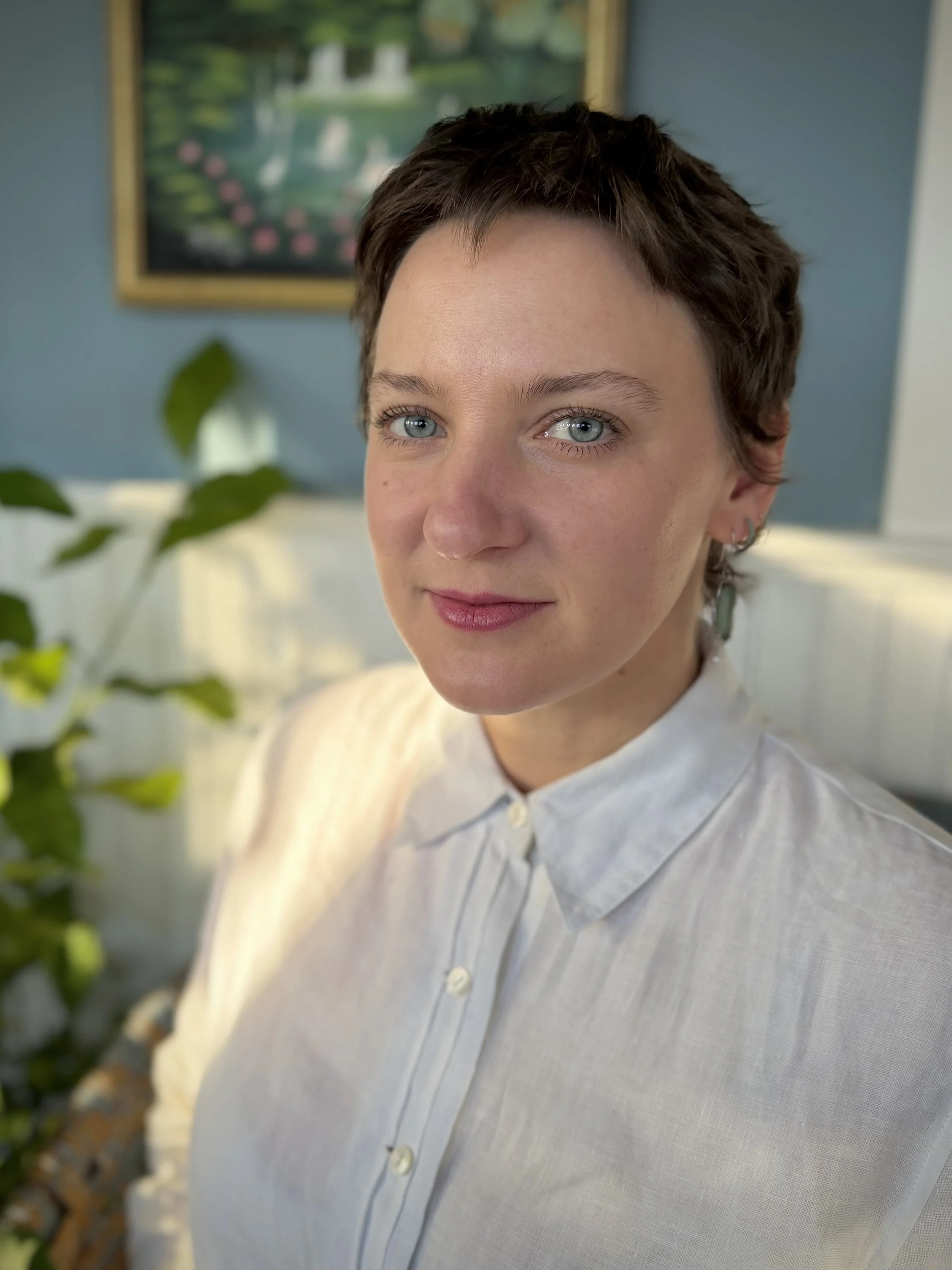 A woman with short dark hair and blue eyes smiles softly, wearing a white button-up shirt, with a framed landscape painting and a green plant in the background.