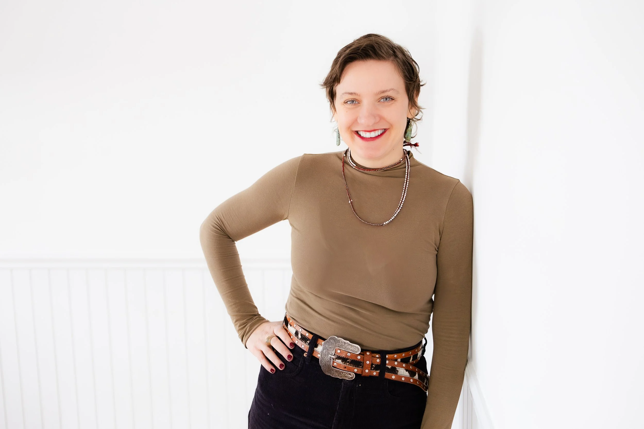 A woman with short brown hair, wearing a long-sleeved tan top, dark jeans, a large belt with a decorative silver buckle, layered necklaces, and earrings, standing against a white background and smiling at the camera.