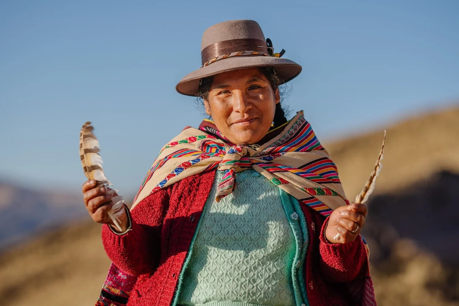 A woman wearing traditional Andean clothing, including a colorful shawl and a hat, holding two feathers outdoors with a mountainous landscape in the background.