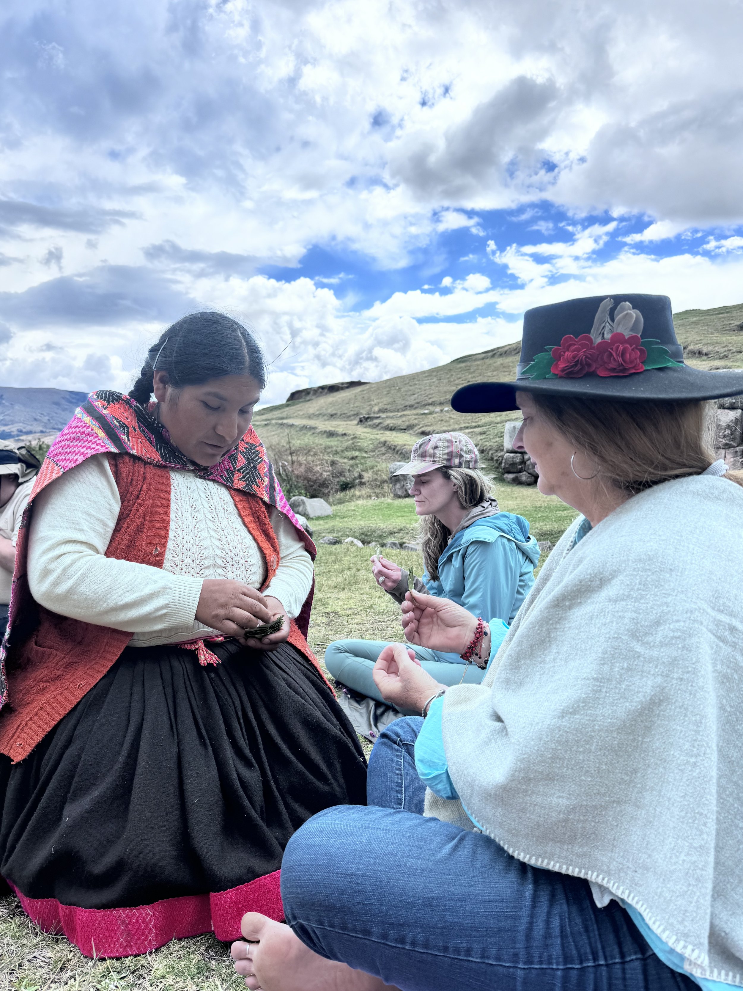 Group of women sitting outdoors on grass, engaging in a spiritual or religious practice, with a scenic landscape of rolling hills and a cloudy sky in the background.