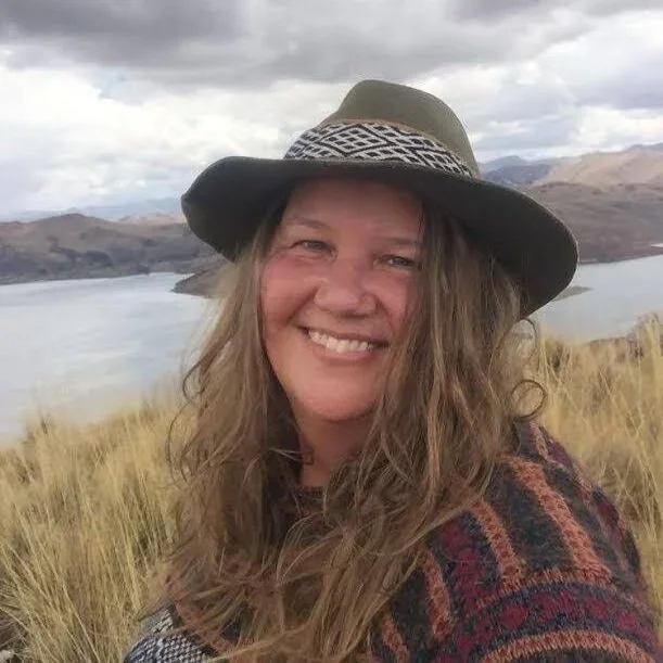 Woman with long wavy hair wearing a wide-brimmed hat and a patterned sweater, smiling outdoors with a lake and mountains in the background.