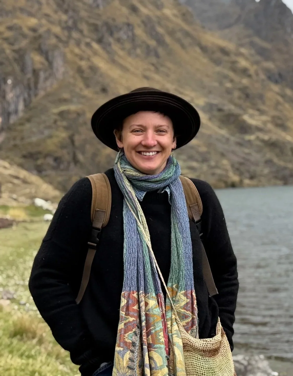 A smiling woman wearing a black hat, multicolored scarves, a black jacket, and a backpack standing outdoors near a body of water with mountains in the background.