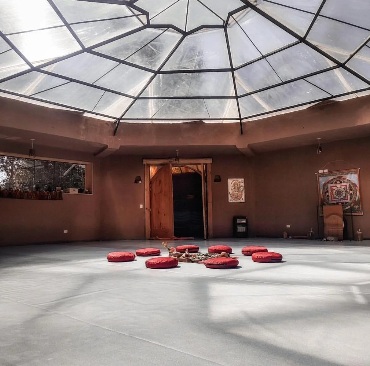 Interior of a round room with a glass dome ceiling, brown walls, a wooden door, and red cushions arranged in a circle on the floor. There are wall hangings and windows providing natural light.