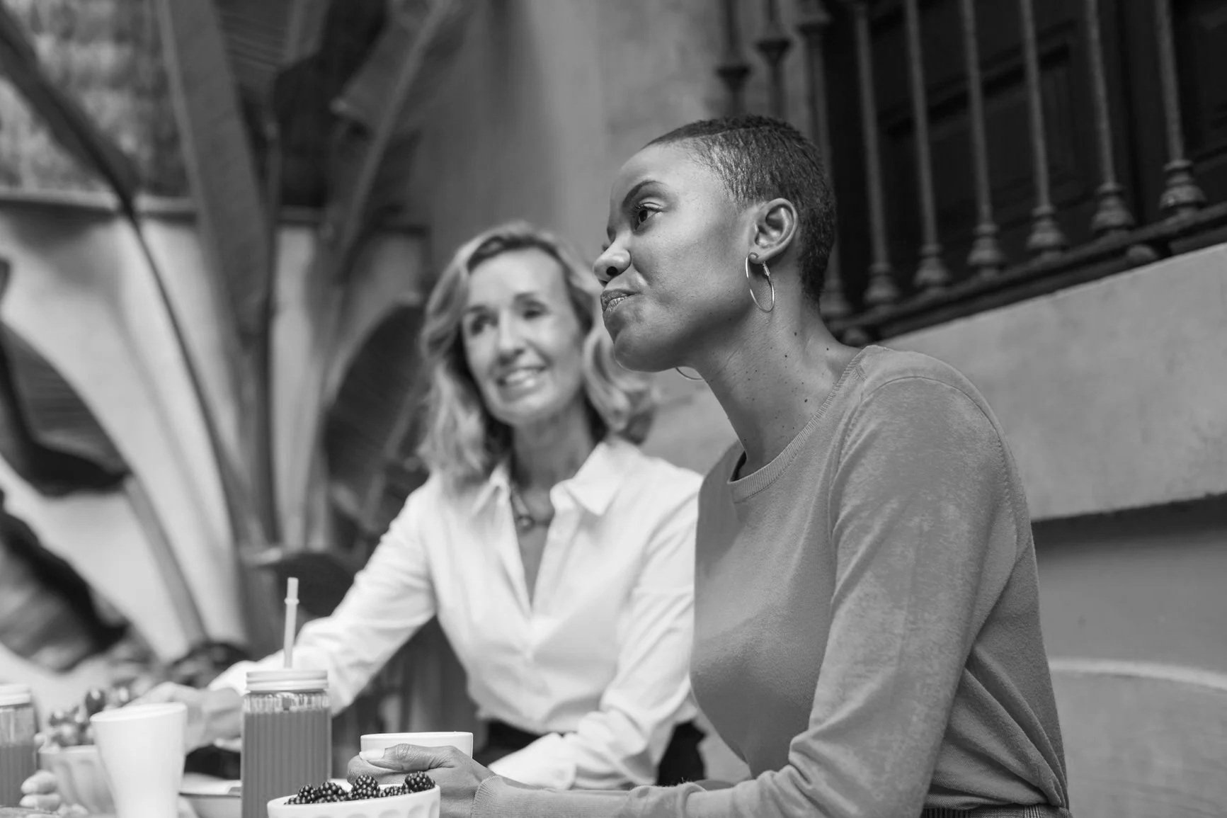Two women sitting at a table with drinks and fruit, engaging in conversation in a cozy, indoor setting with a staircase railing in the background.