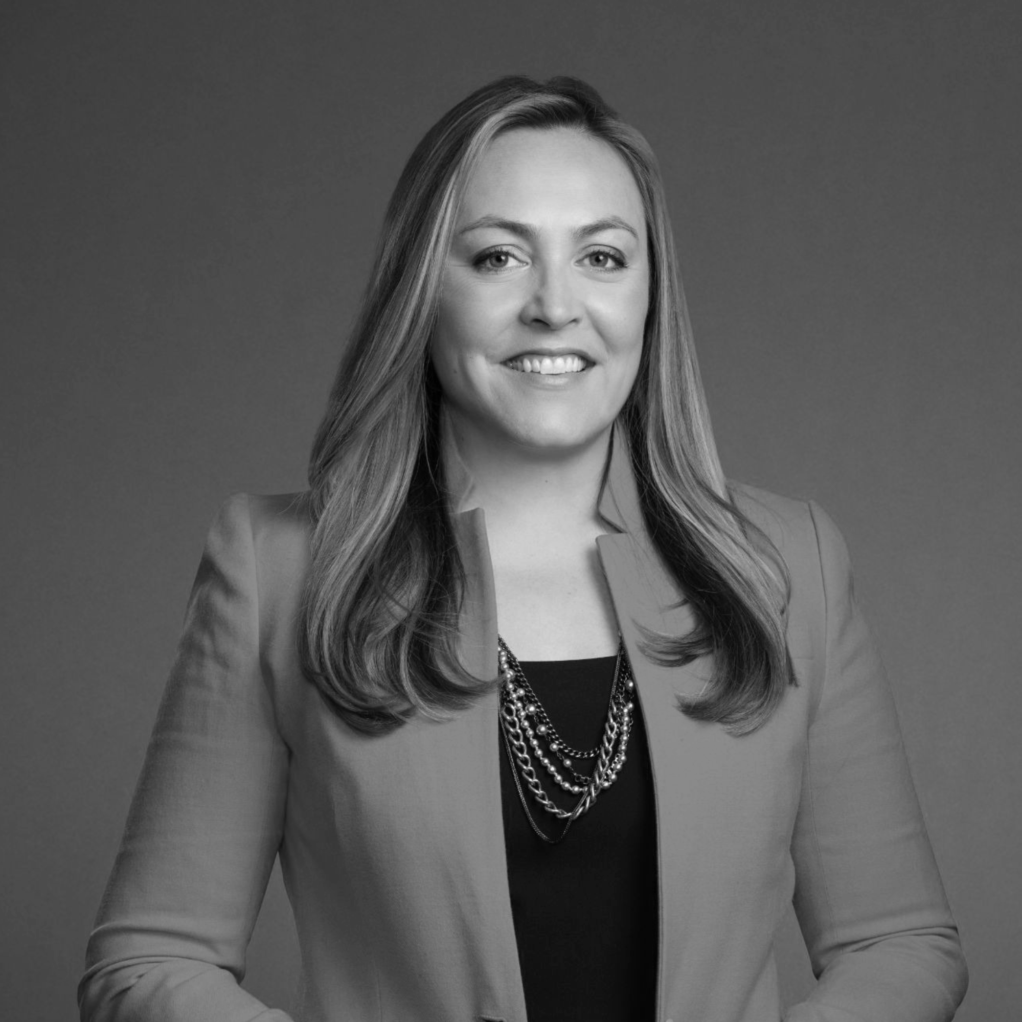 Black-and-white portrait of a smiling woman with shoulder-length hair, wearing a blazer, a top, and layered pearl necklaces, posing against a plain background.