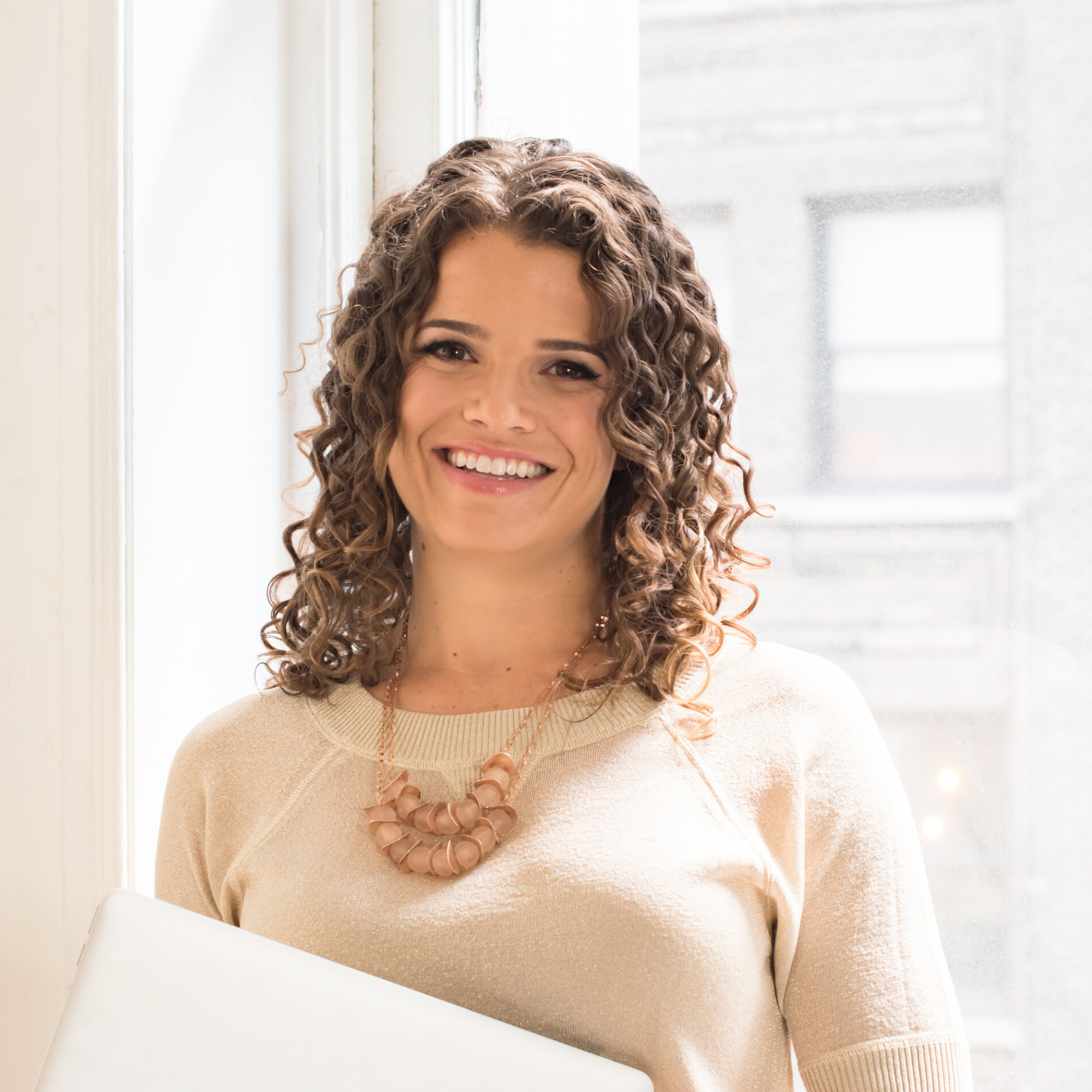 Smiling woman with curly hair wearing a beige top and chunky necklace, sitting by a window.