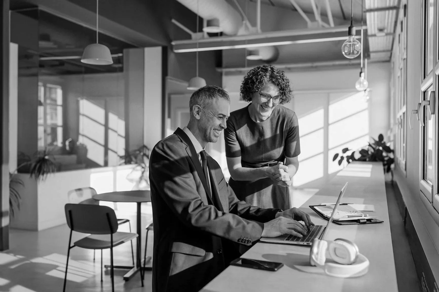 A man in a suit and a woman with curly hair and glasses are sharing a laugh while looking at a laptop in a modern office space with large windows and hanging lights.